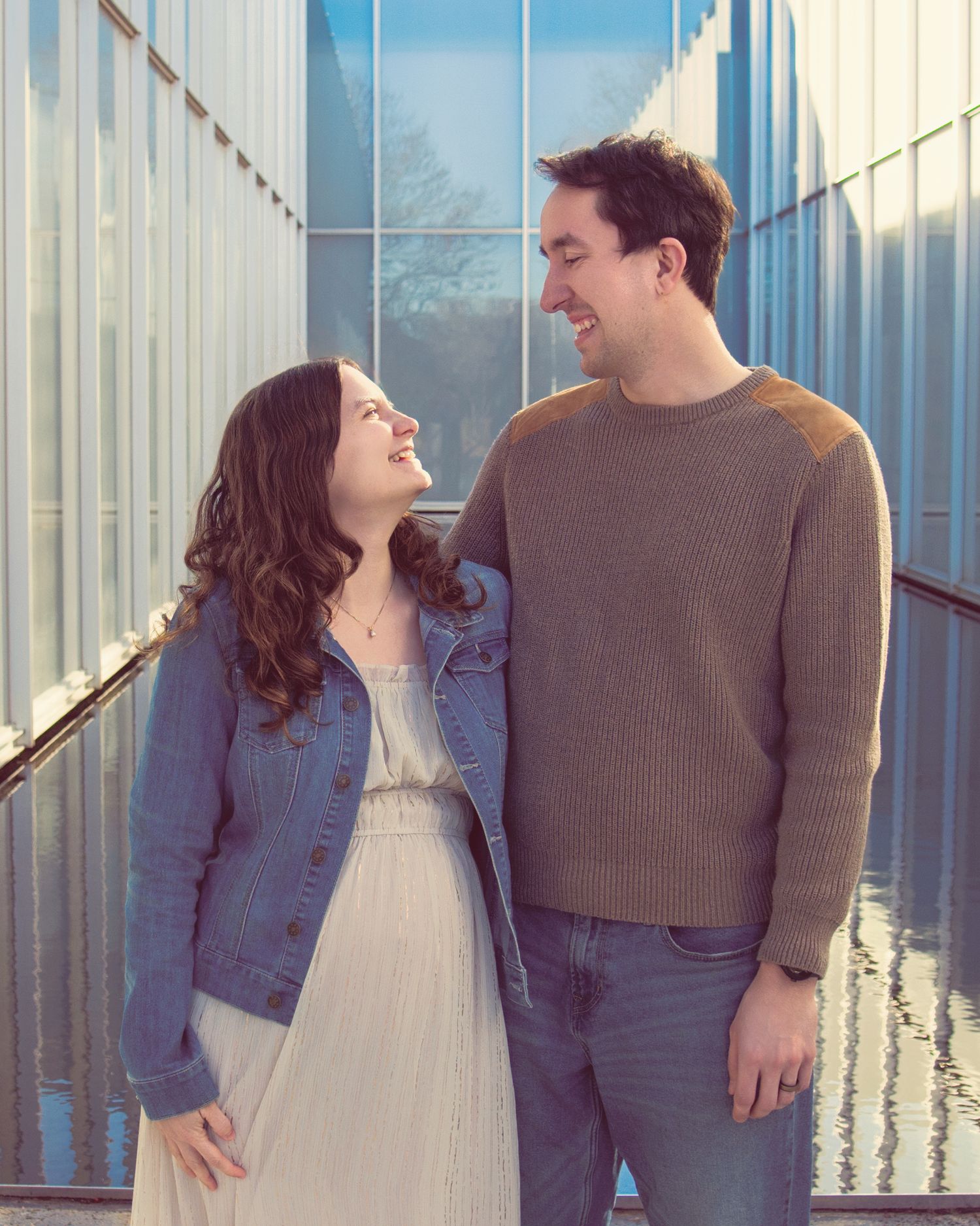 A couple stands together in a bright glass-lined hallway, smiling at each other in a sweet candid moment.