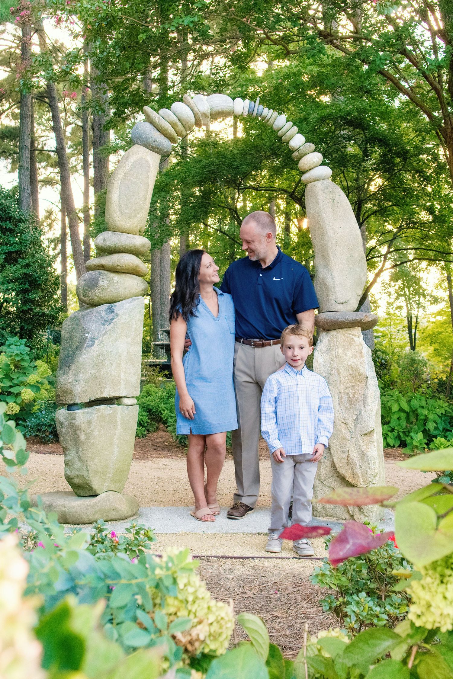 A family stands together by unique stone sculptures in a garden path surrounded by lush greenery and flowering plants.