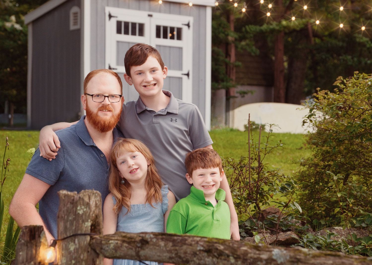 Outdoor family portrait taken in front of a gray shed with string lights in the background.