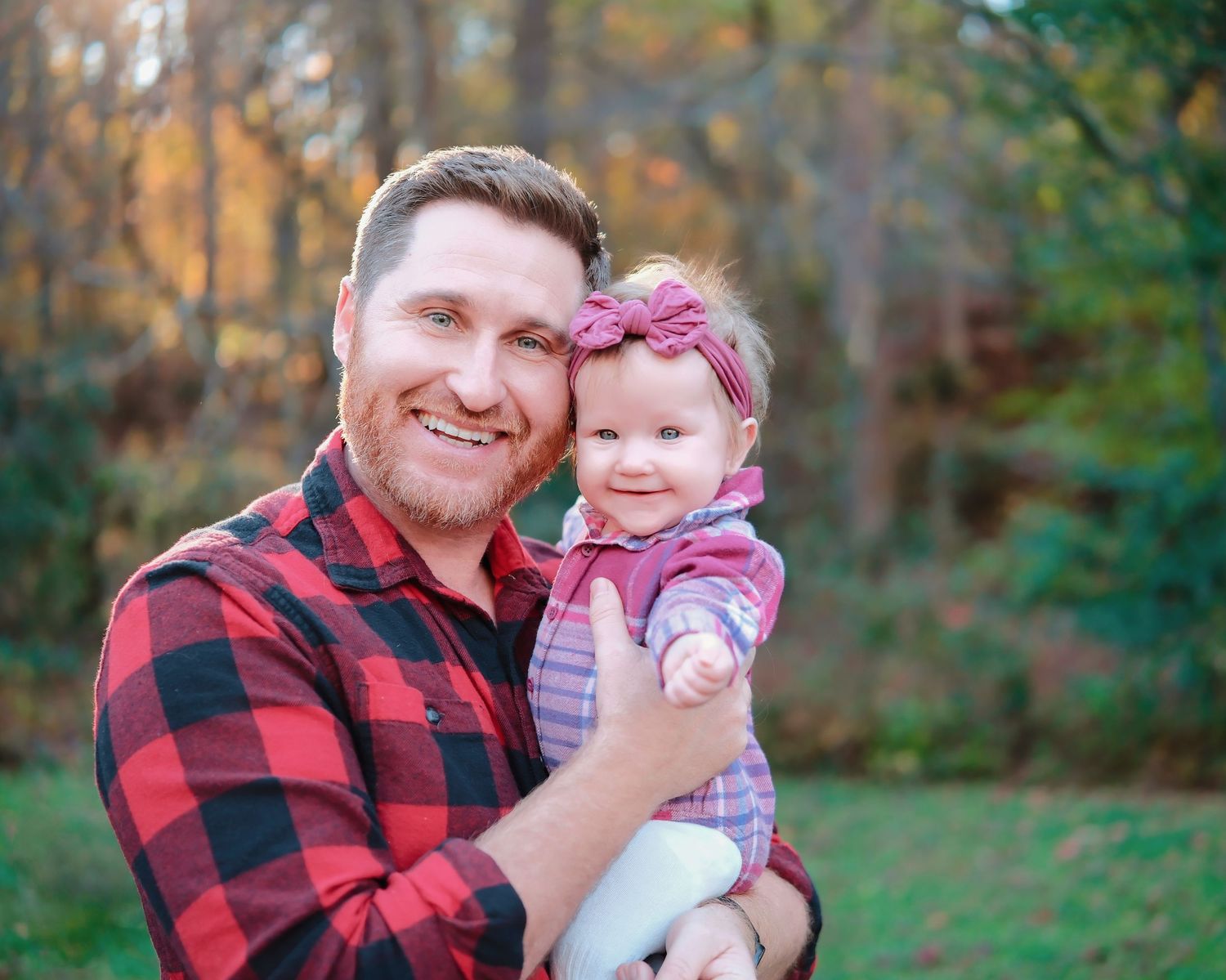 A person in a red plaid flannel shirt holds a small baby wearing a pink headband outdoors in autumn.