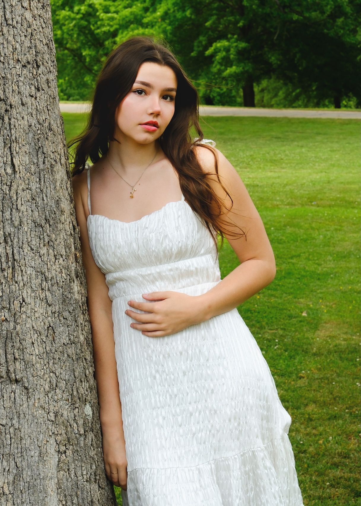 Person in white summer dress poses against tree in sunny park setting.