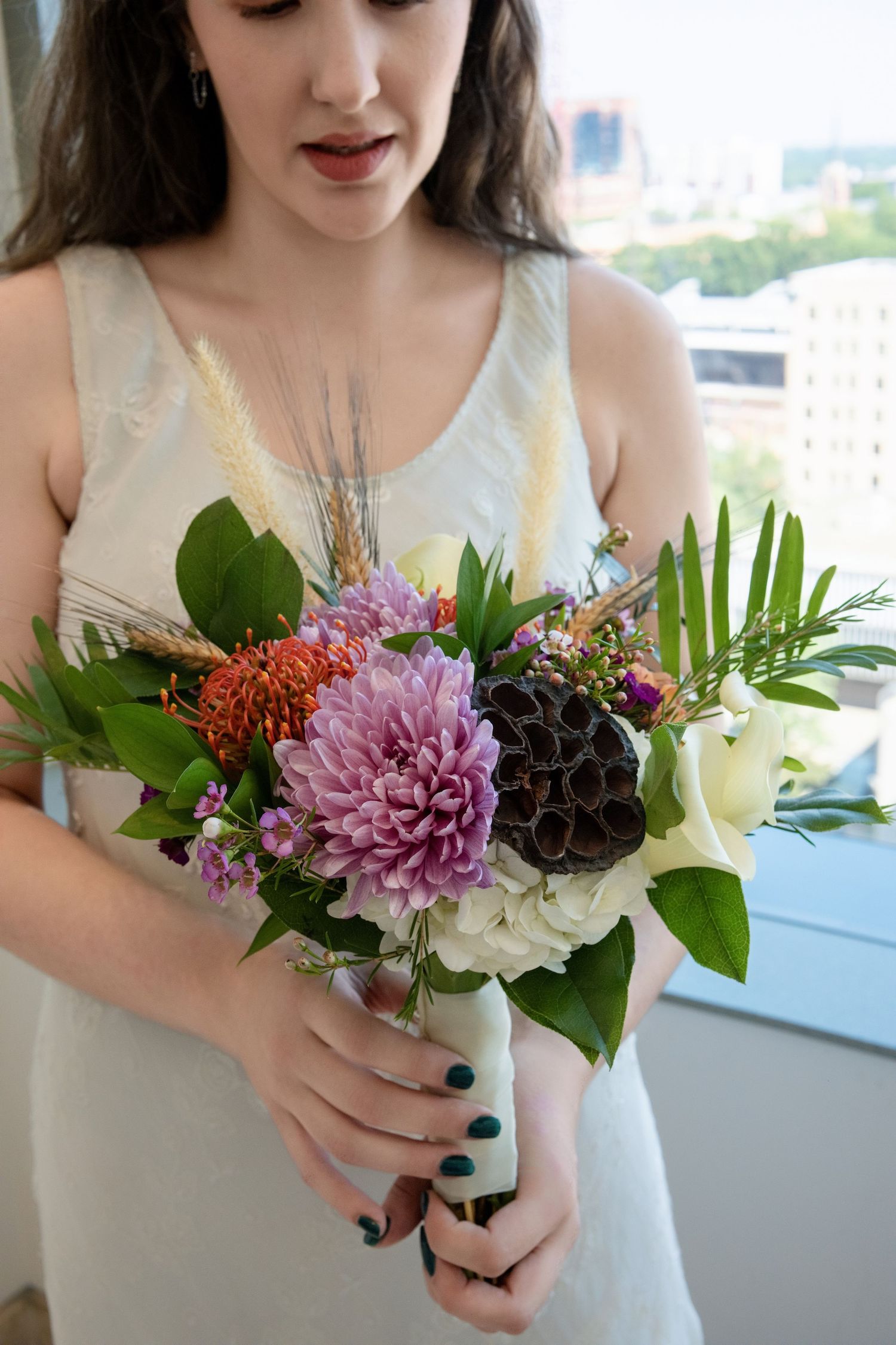 A bridal bouquet featuring pink dahlias, white roses, and tropical greenery held against a white dress with city views behind.