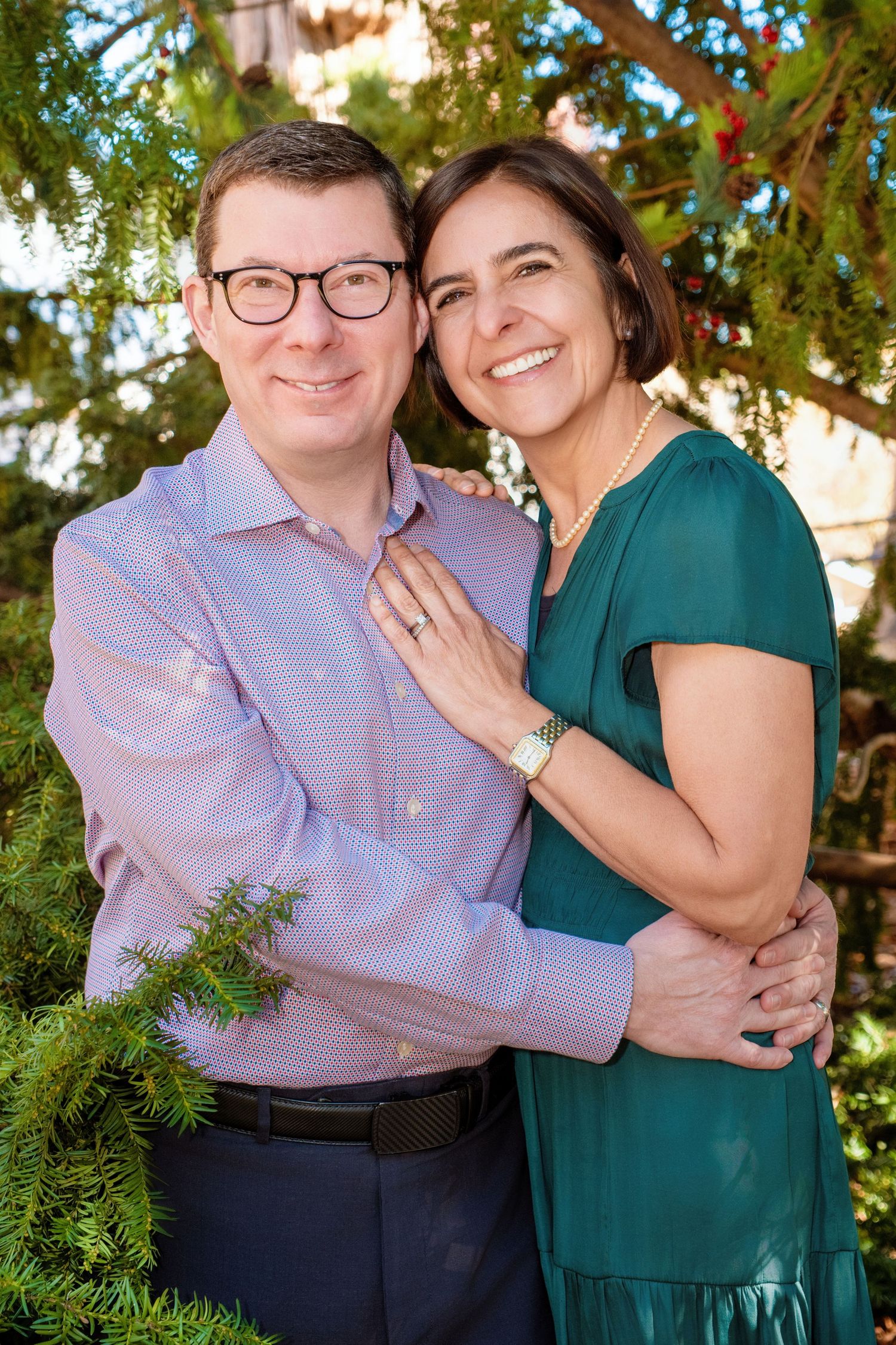 A couple in green and purple attire share intimate moments while posing outdoors near evergreen foliage.