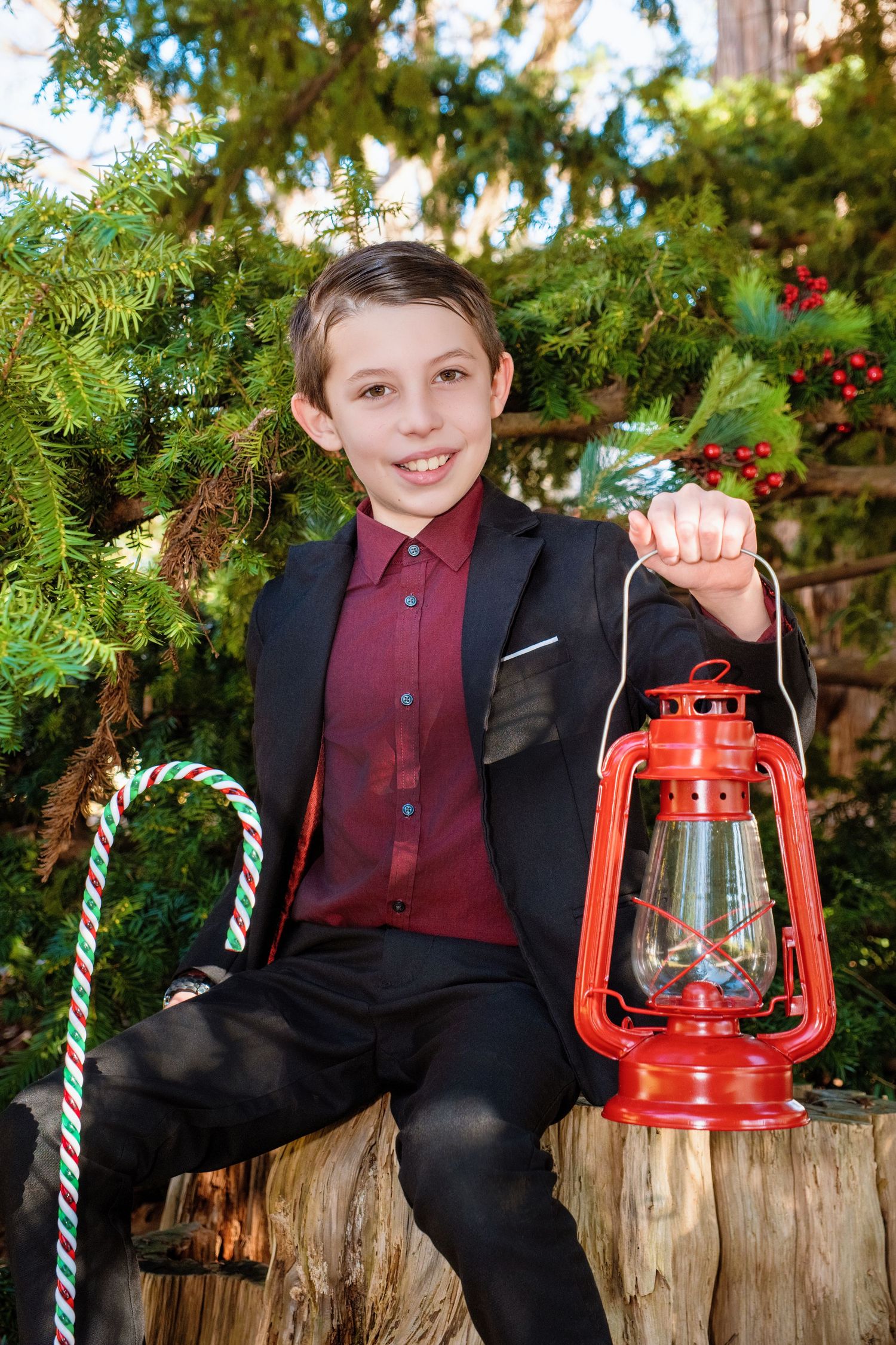 Child in black suit and burgundy shirt holds red lantern while sitting on tree stump during holiday photo session.