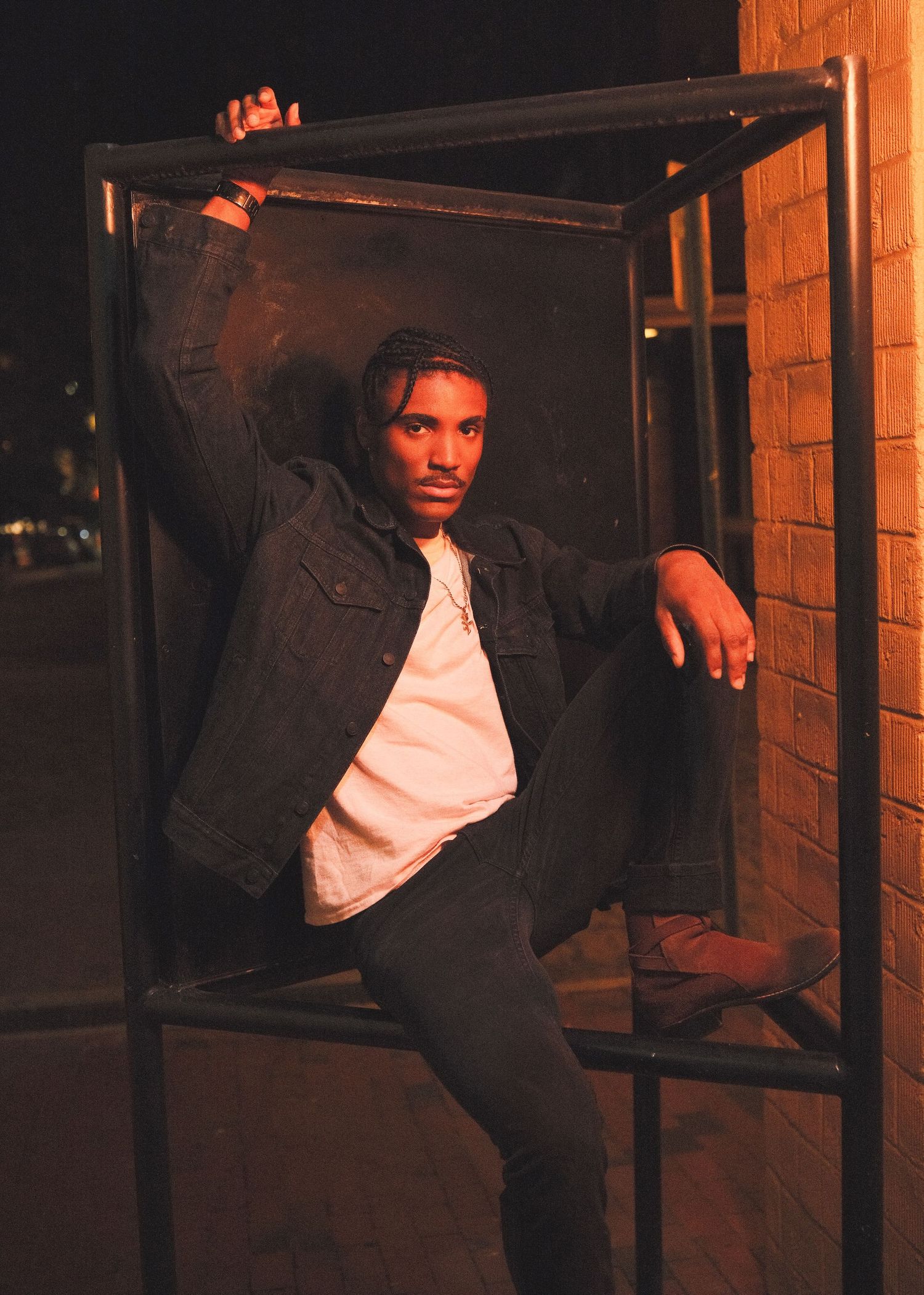 Person in dark jacket posing against brick wall at night in moody lighting.