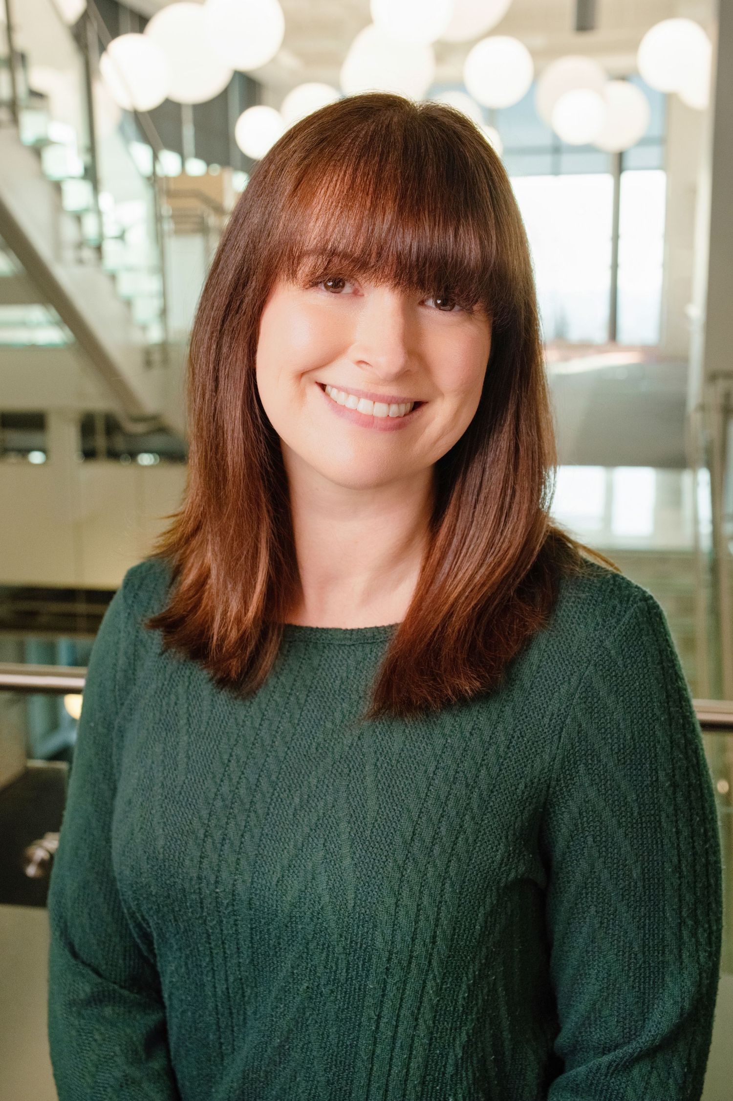 Professional headshot of person in green sweater with straight brown hair against bright modern interior.