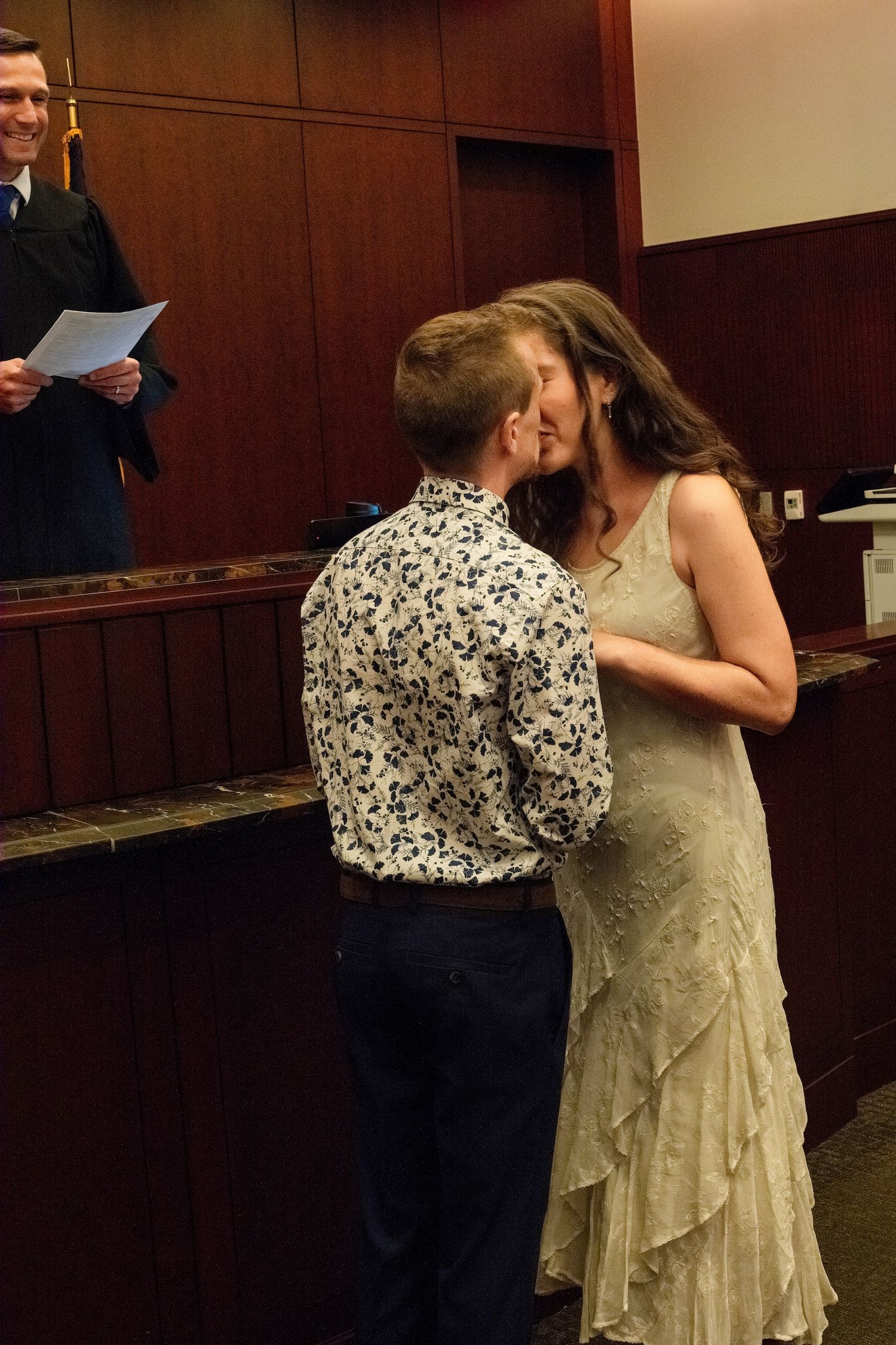 A couple shares their first kiss at a courthouse wedding ceremony with an officiant standing nearby.
