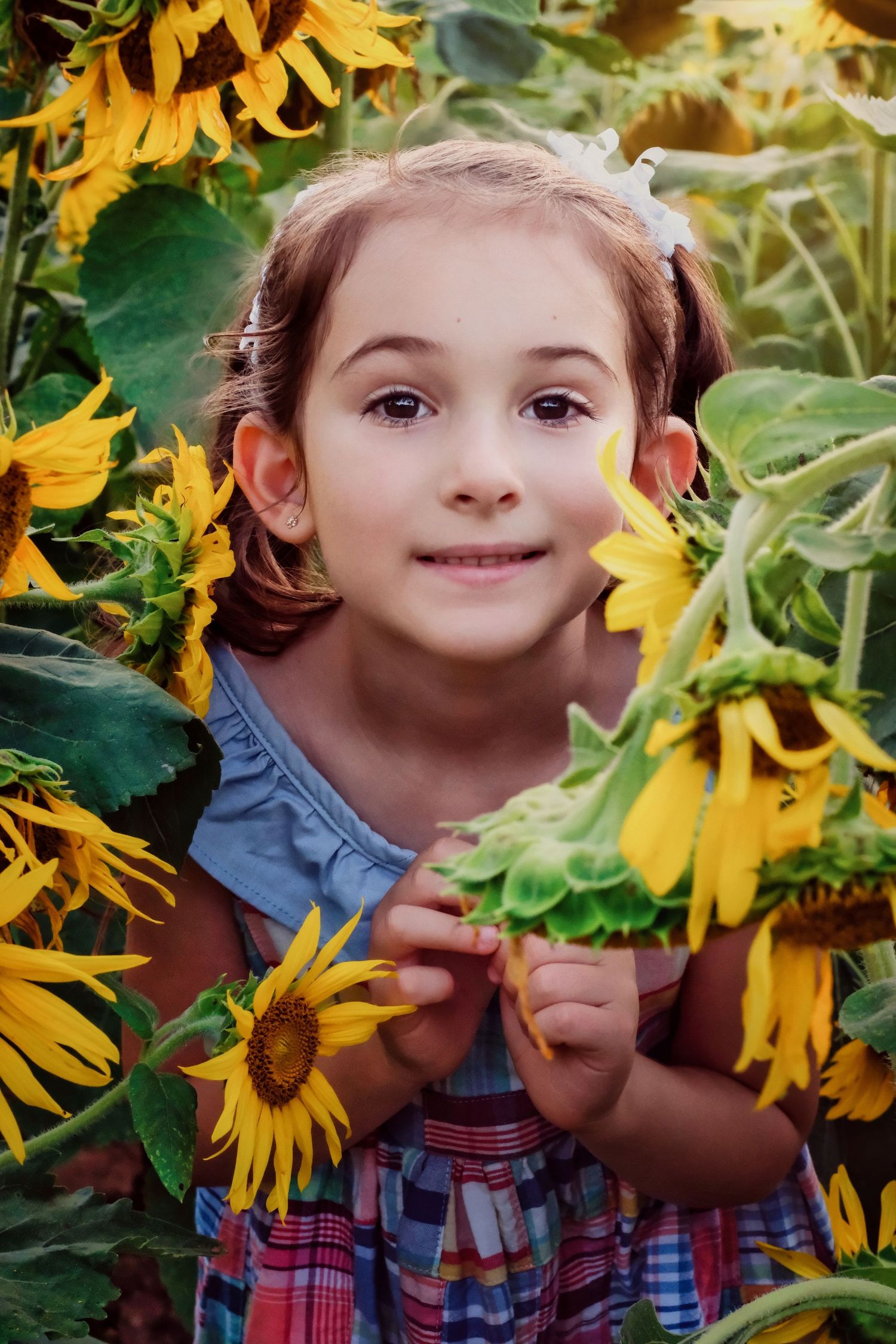 Young girl in plaid dress poses among bright yellow sunflowers in garden setting.