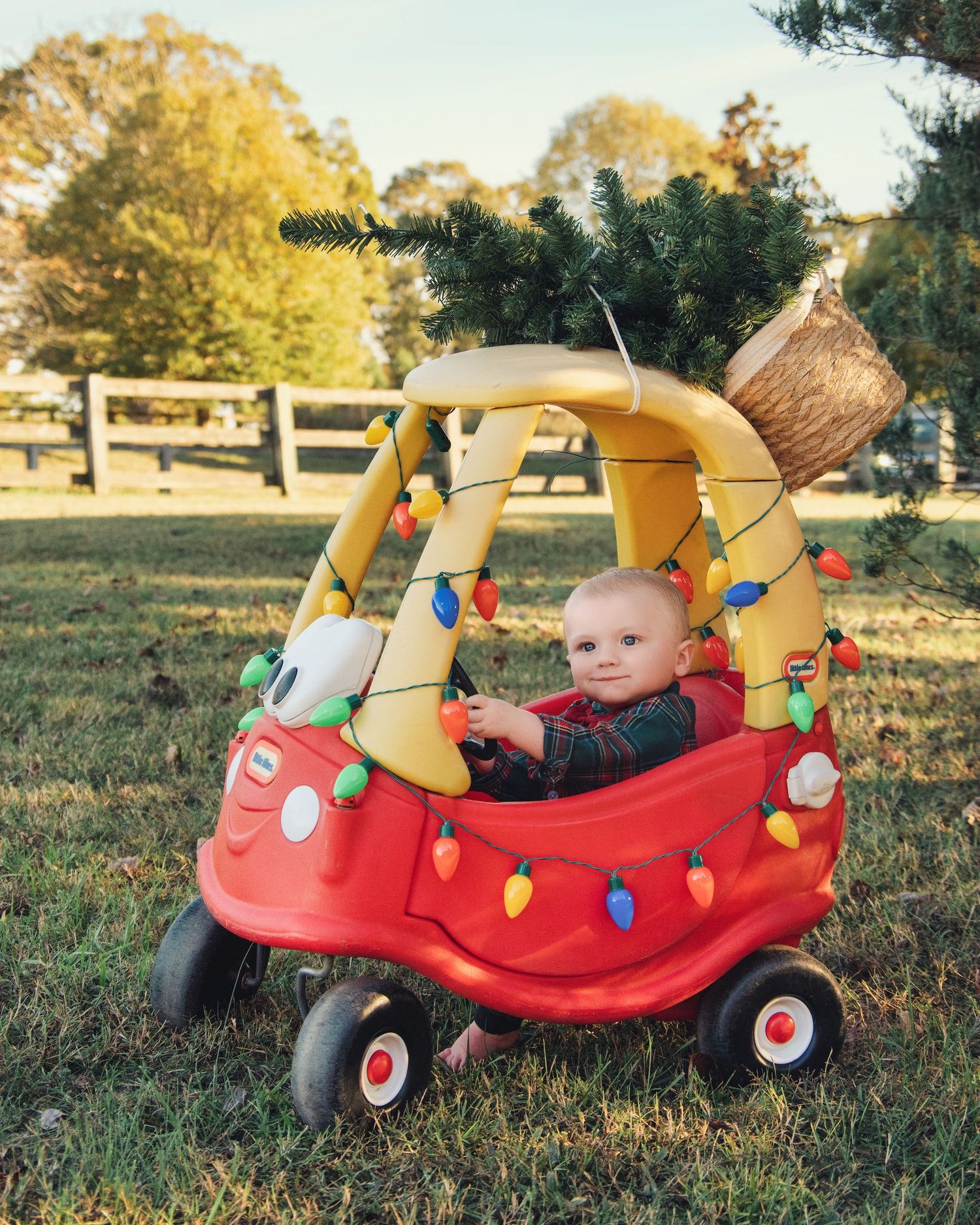 A red and yellow toy cozy coupe car decorated with festive holiday lights sits on grass near a wooden fence.