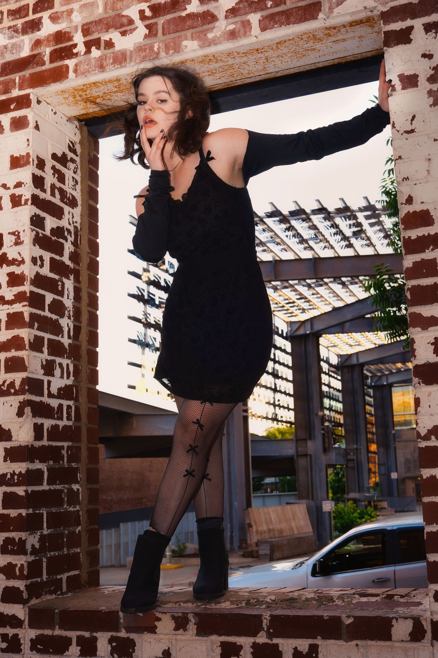 Dynamic fashion pose in black dress framed by brick archway with urban scenery.