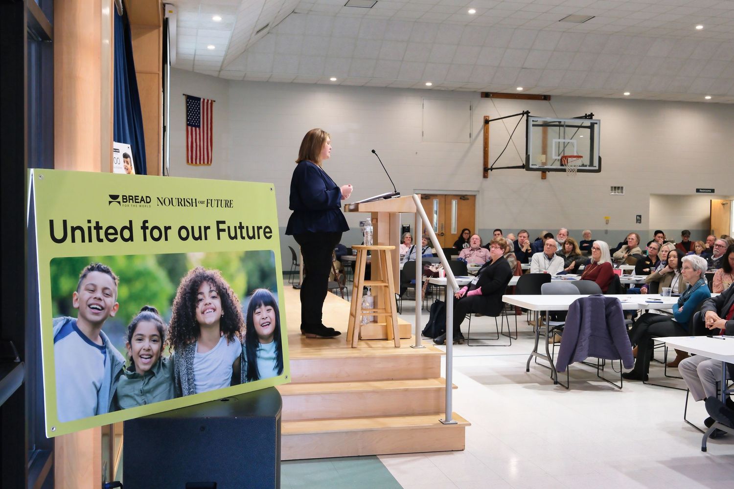 A speaker addresses a crowd from a podium in a gymnasium with a United banner displayed in the foreground.