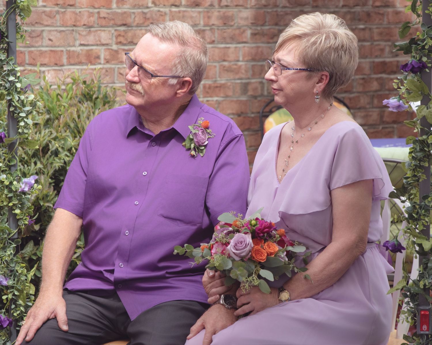 A couple in purple and lavender attire sits together in a garden setting with floral bouquet and brick wall backdrop.