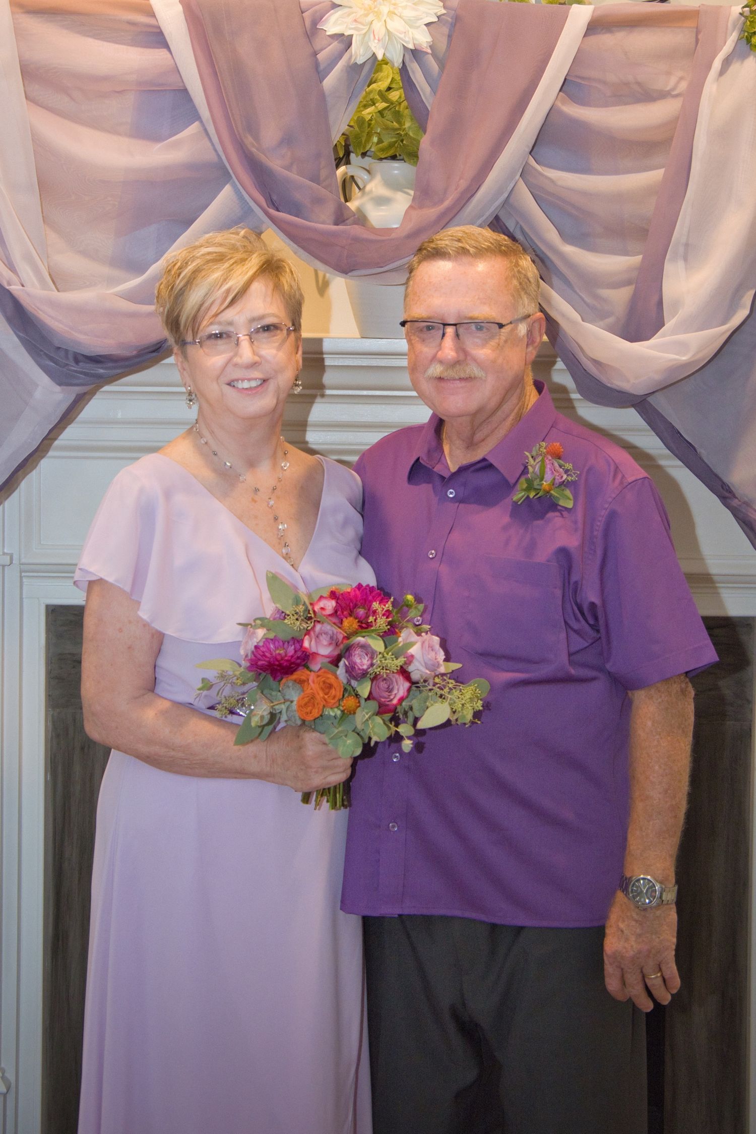 A formal couple poses at a wedding event, wearing coordinating purple attire with a floral bouquet and decorative backdrop.