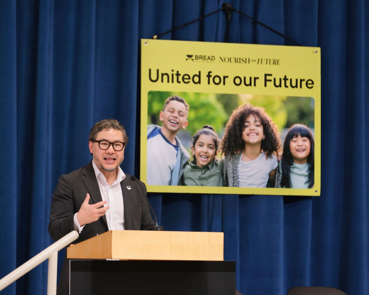 A presenter stands at a podium in front of a display banner showing a group photo with text 'United for our Future'.