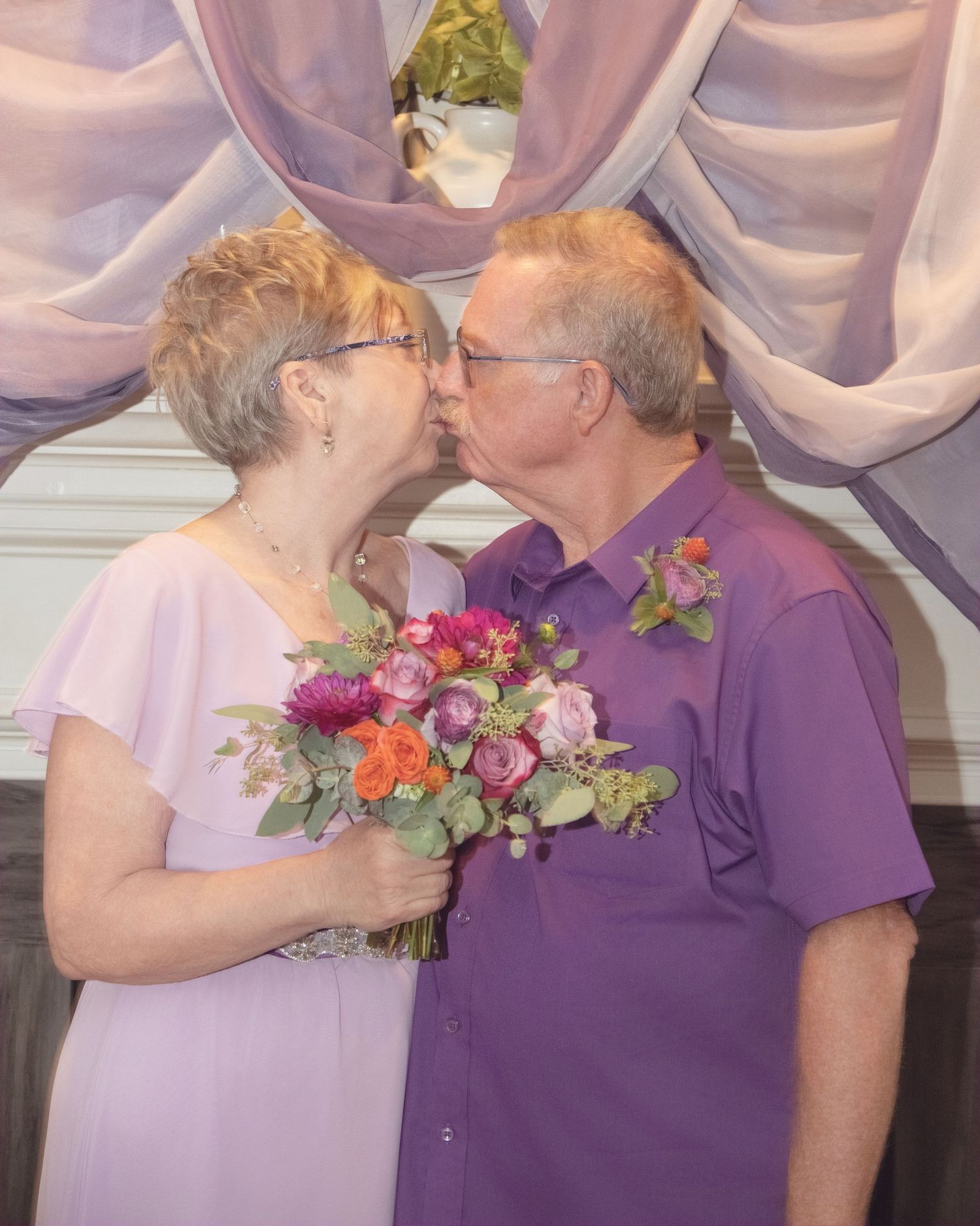 A couple in matching purple attire share a tender moment with a pink and orange floral bouquet at a decorated venue.