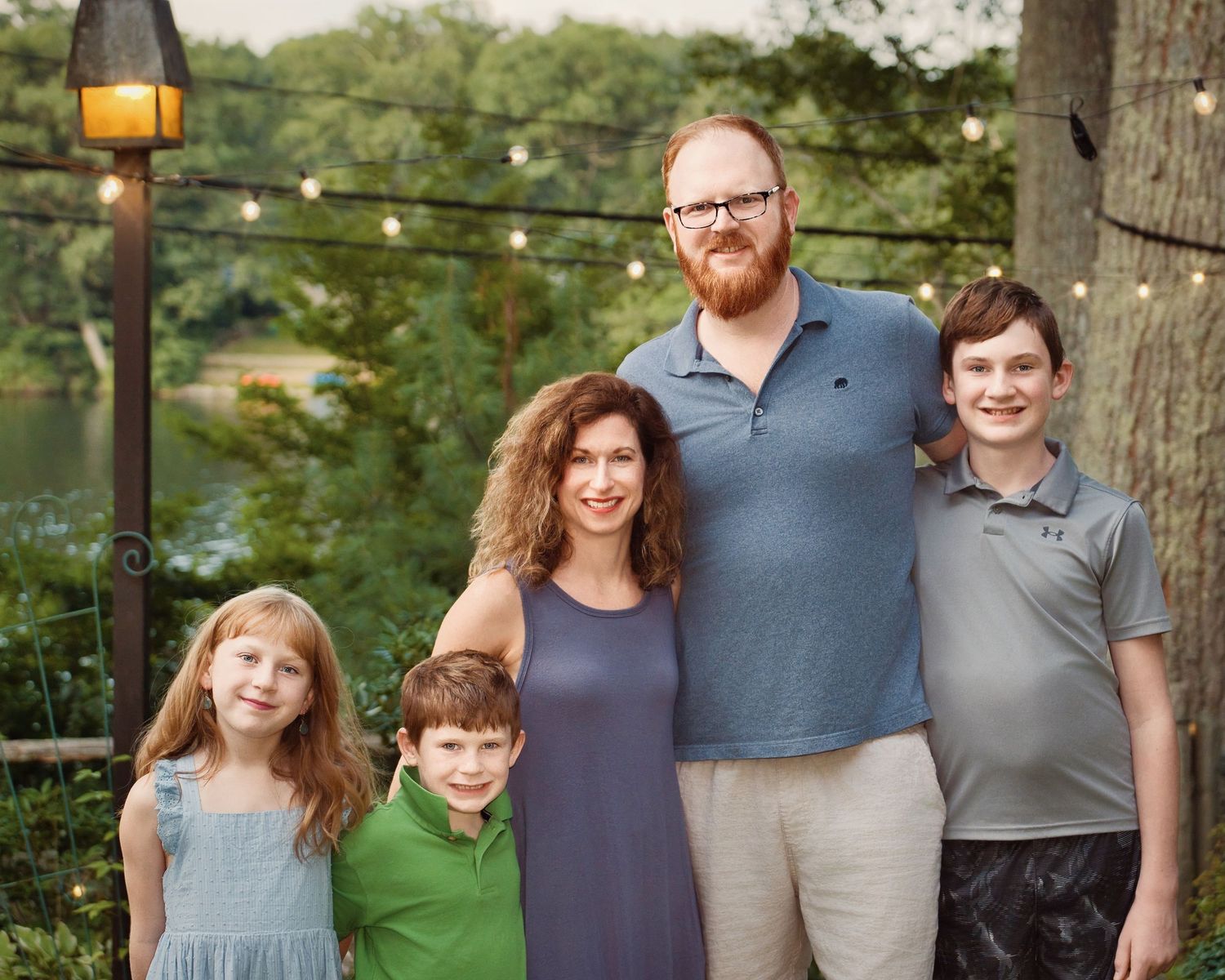 Family of five poses outdoors at dusk with string lights in the background.