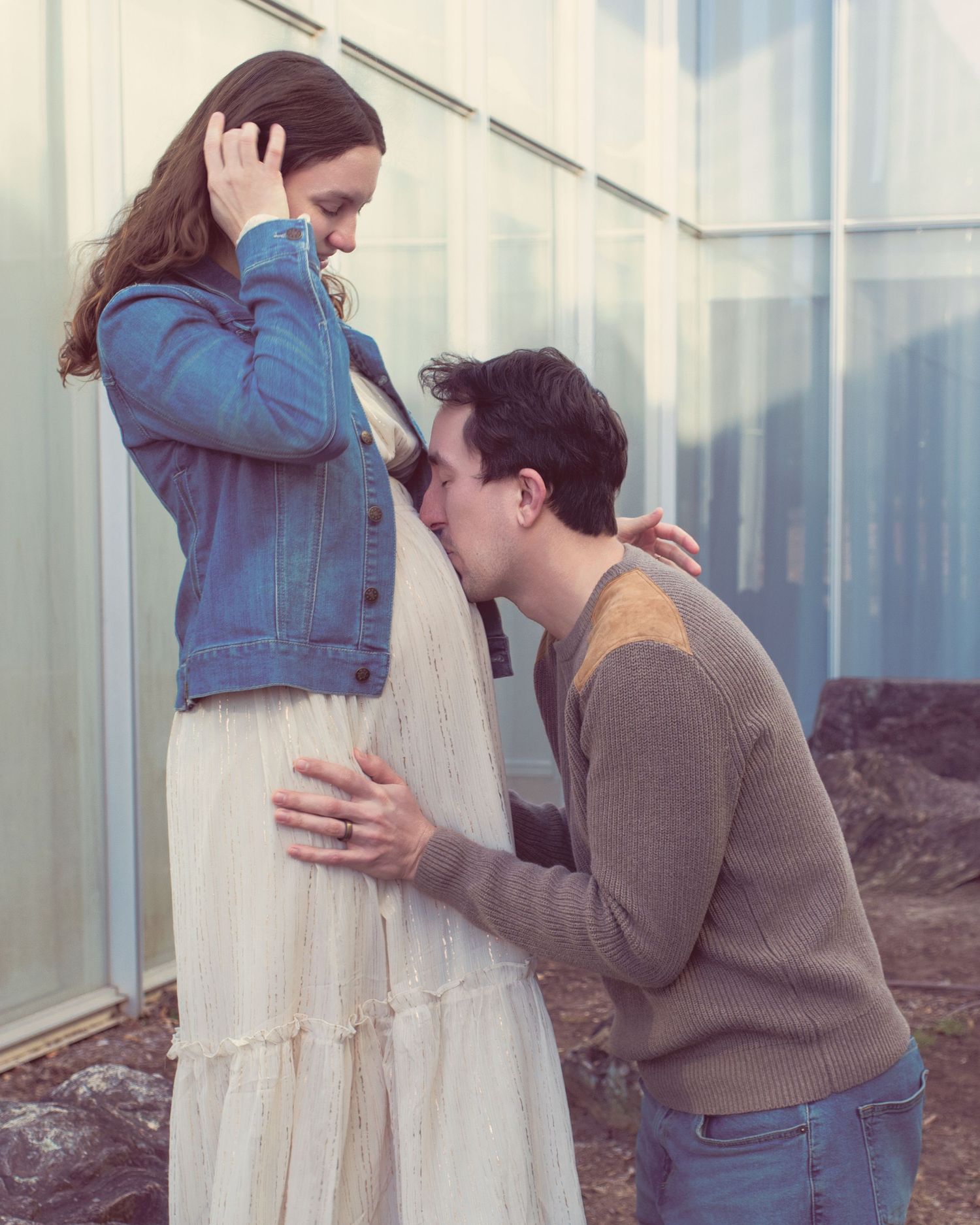 A tender romantic moment between two people in casual attire standing beside a wall in natural lighting.