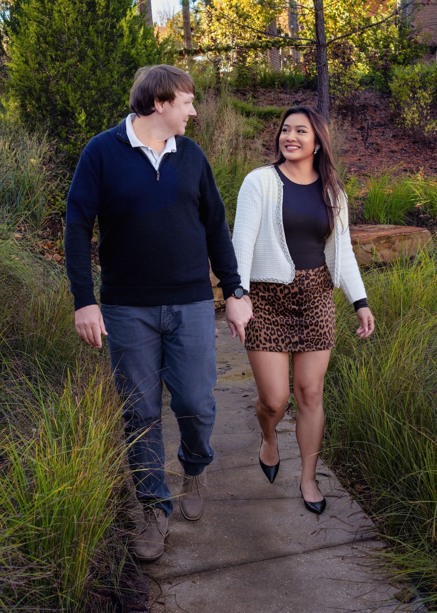 A couple walks hand in hand on a garden path during autumn, wearing casual outfits in a natural outdoor setting.