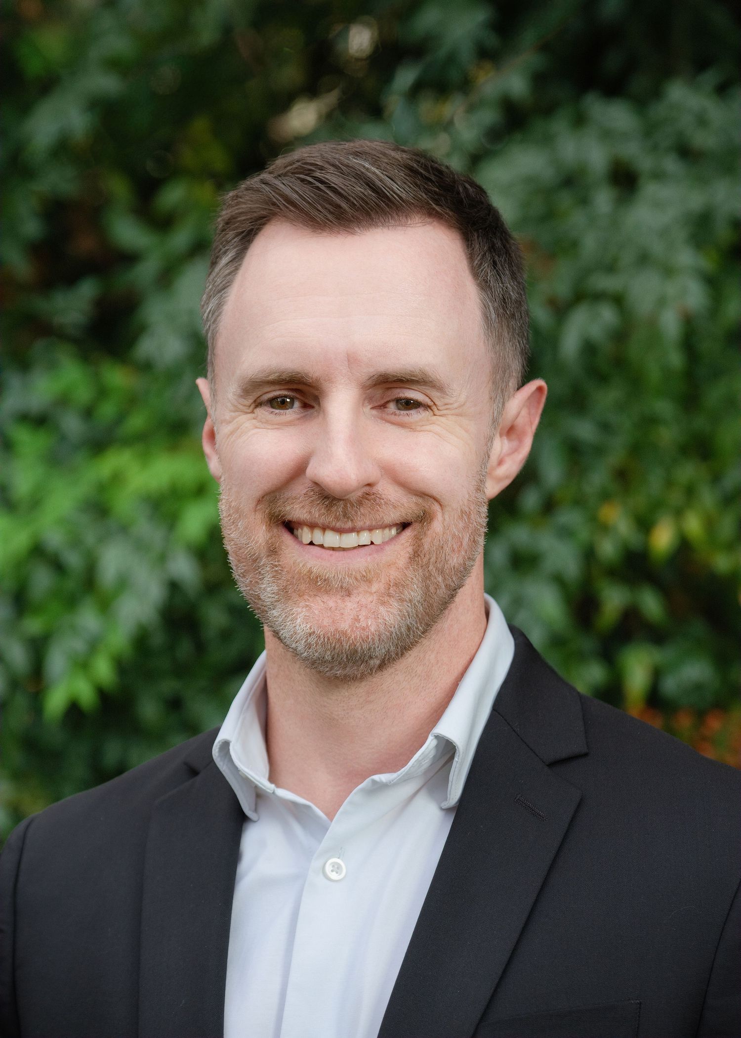 Professional headshot of person in black suit jacket and white dress shirt smiling outdoors with greenery background.