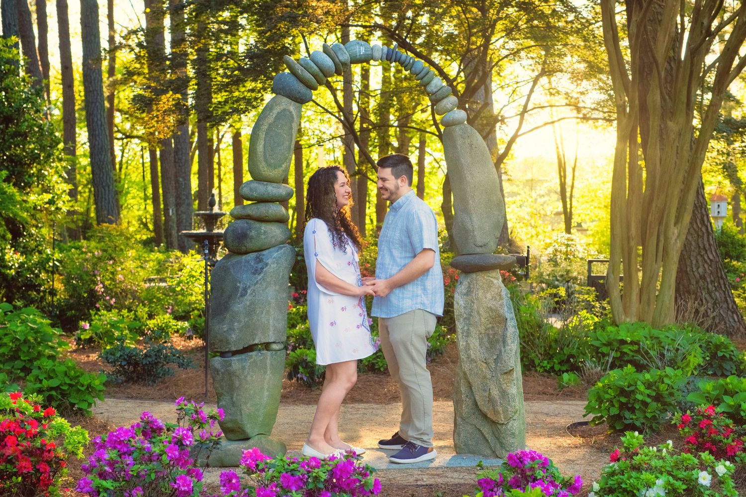 Two people stand under a stone archway surrounded by colorful flowers and trees in a garden at sunset.