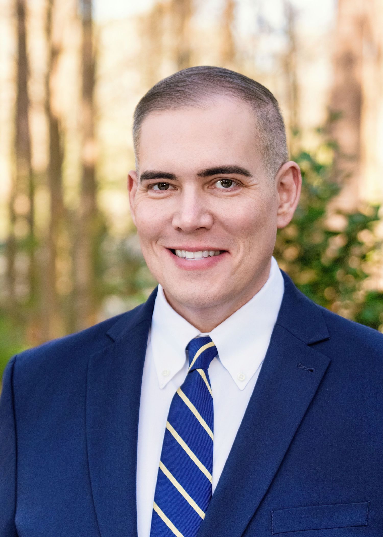 Professional headshot of someone in a navy suit and striped tie smiling at the camera against a blurred outdoor background.