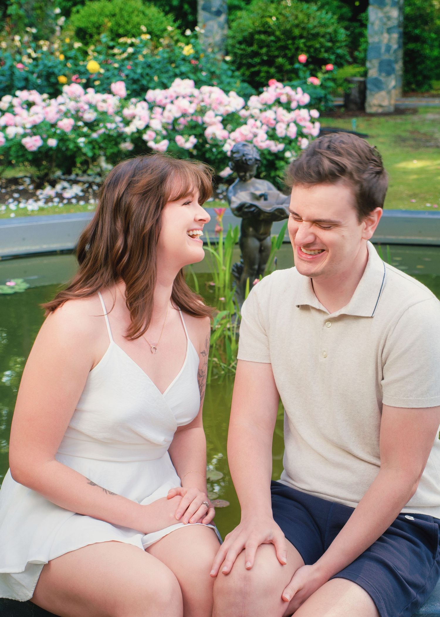 Two people share a tender moment on a park bench surrounded by blooming pink roses in a garden setting.