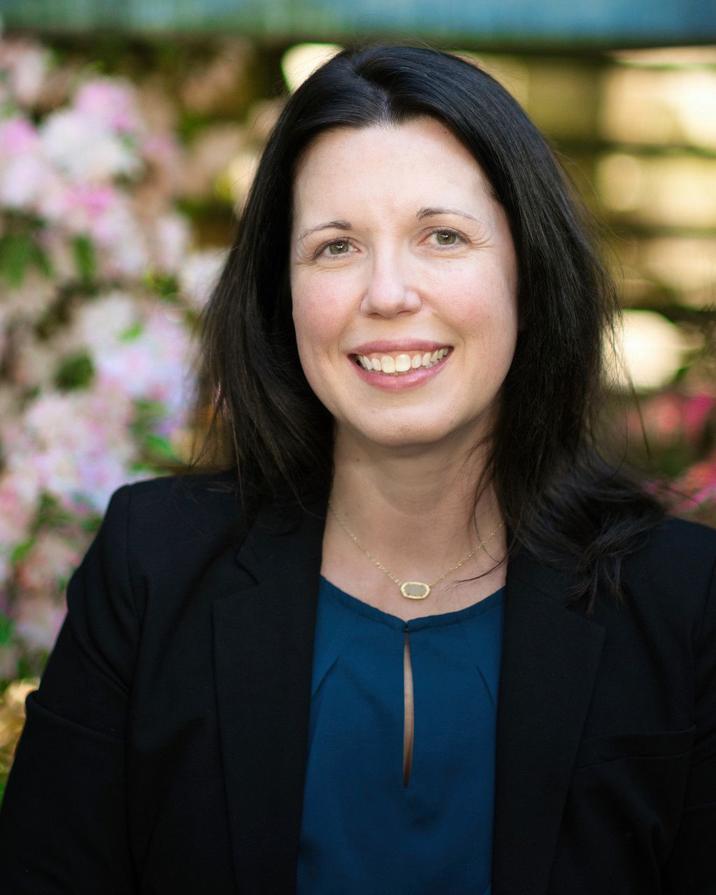 Professional headshot of person in black blazer and navy blouse smiling with pink flowers in background.