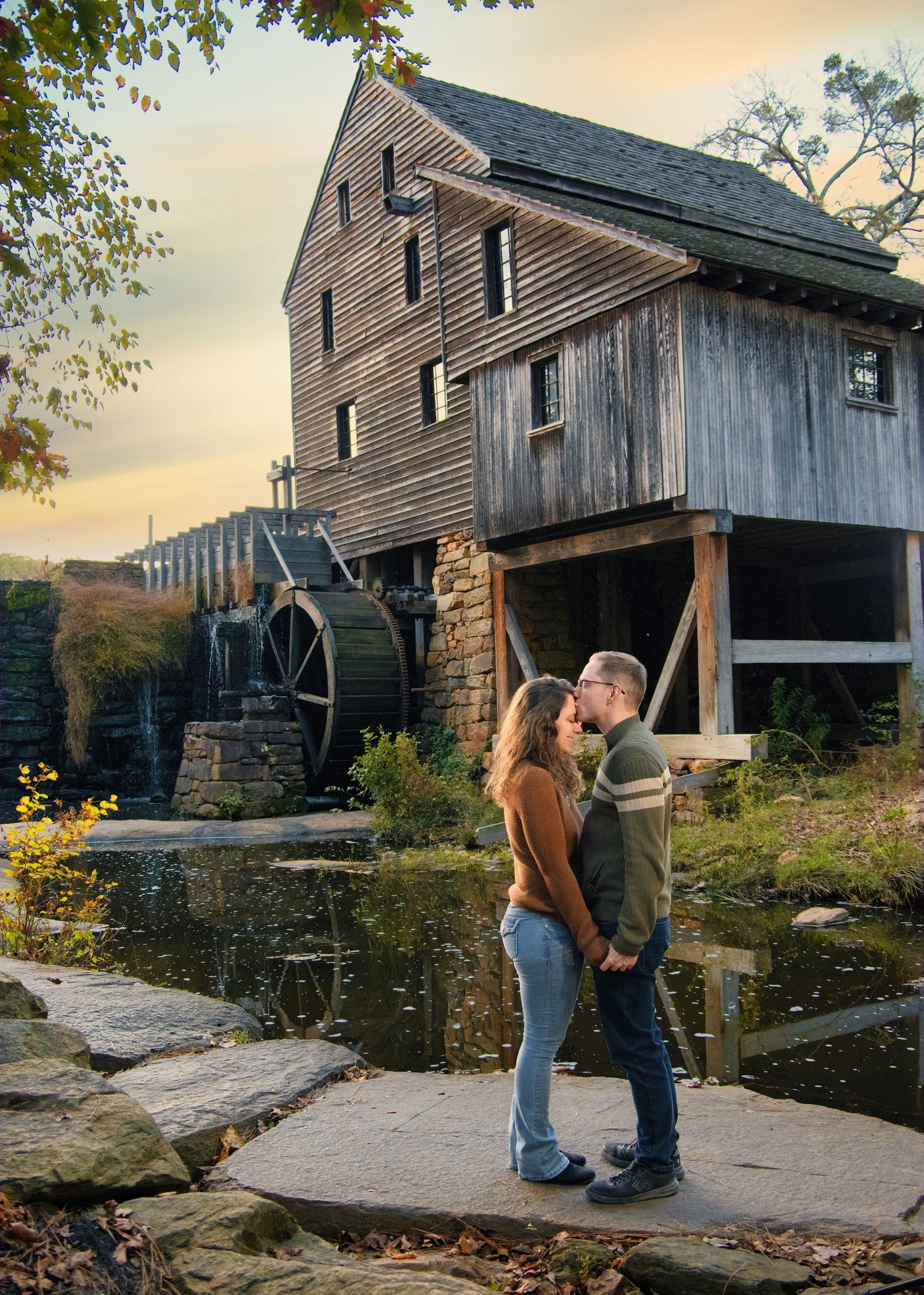 A couple shares a romantic moment in front of a historic water mill at sunset.