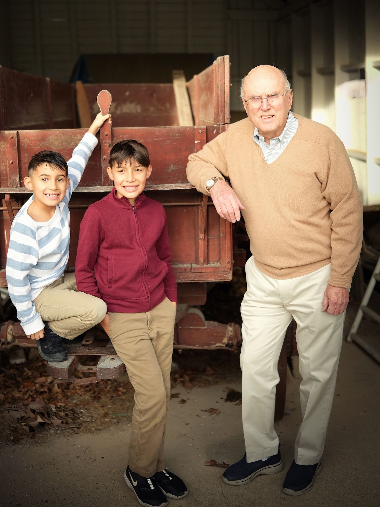 Three people pose by an antique red wagon in a rustic indoor setting.
