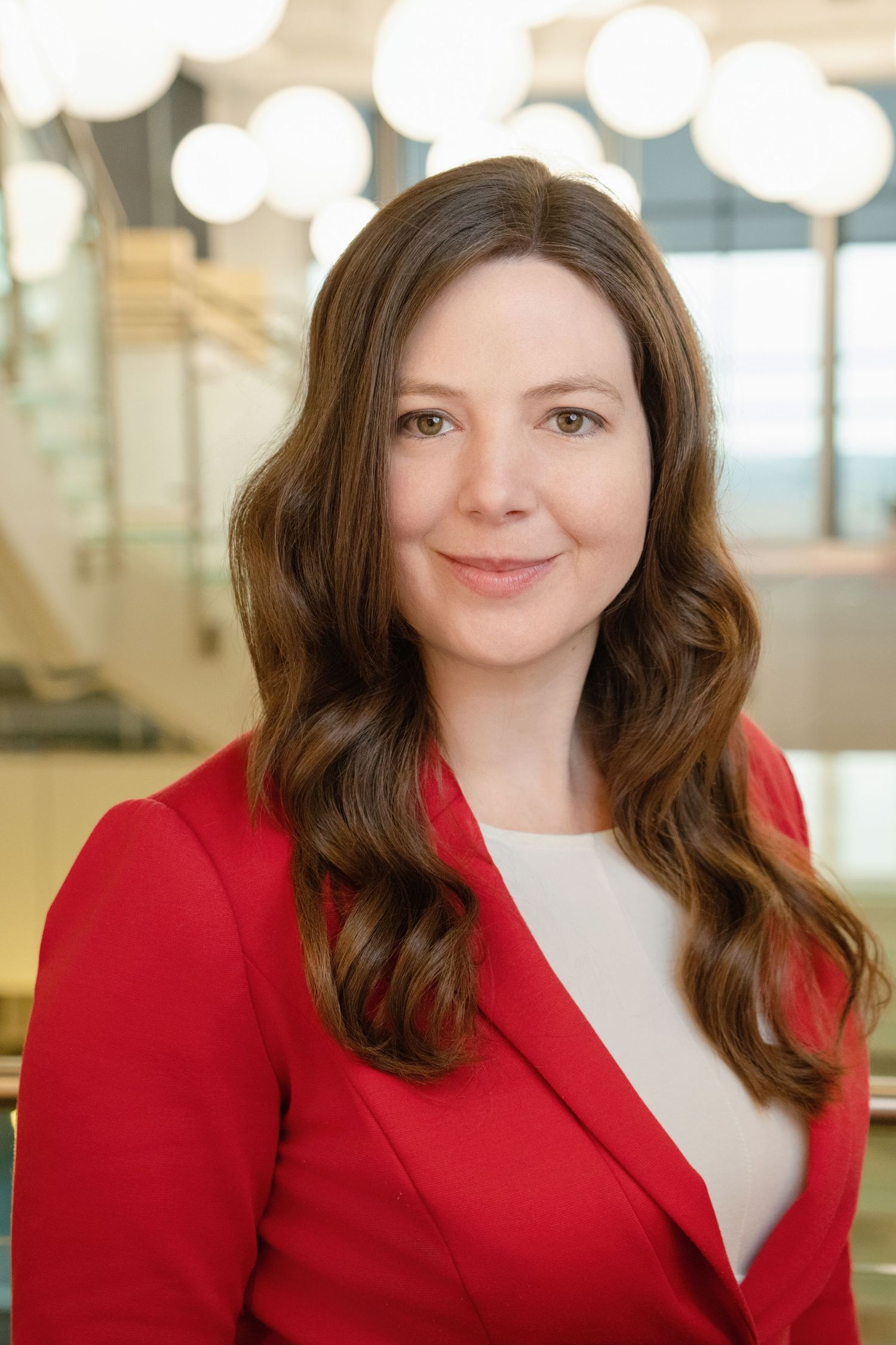 Professional headshot of someone wearing a red blazer and white top with long brown wavy hair in an office setting.