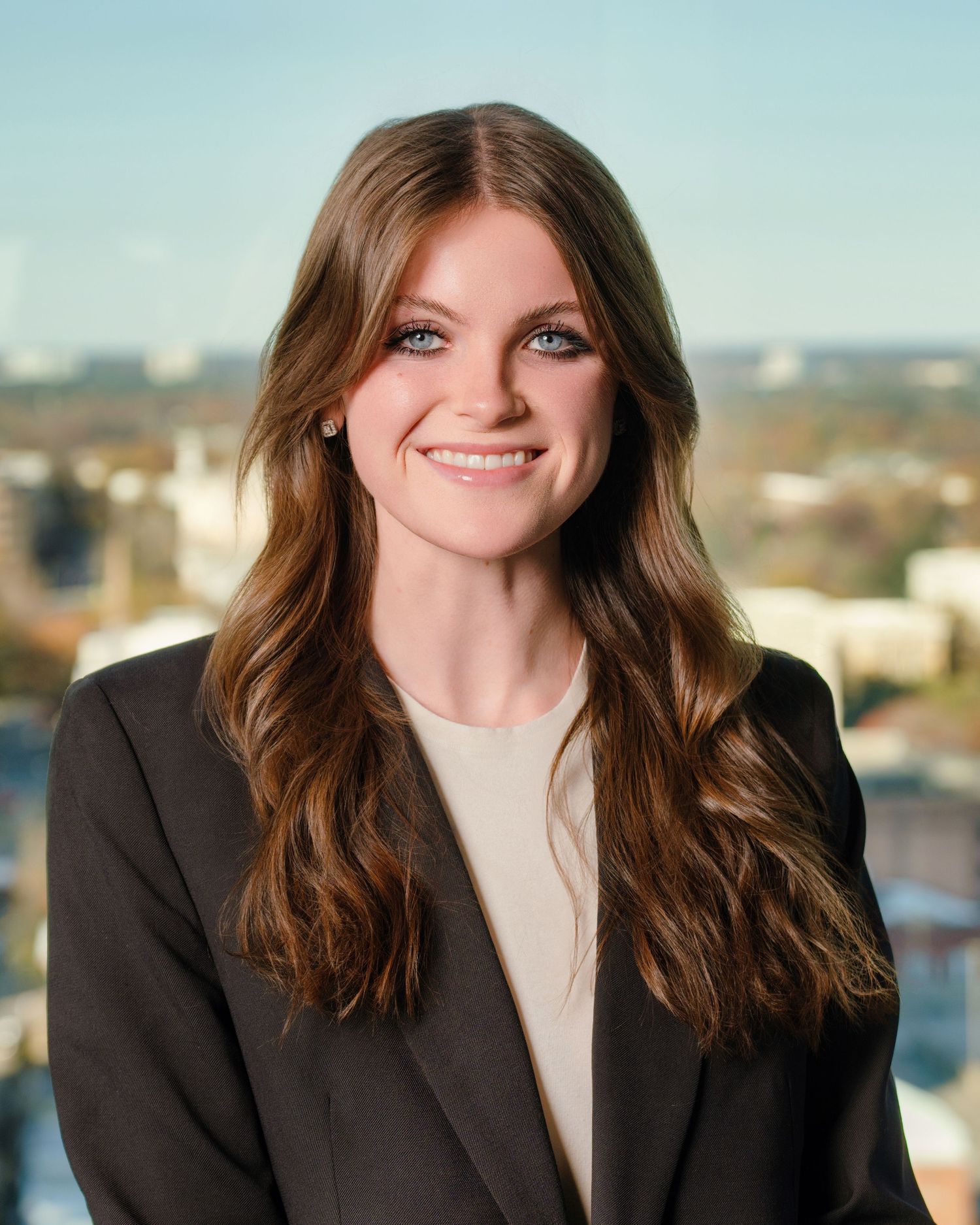 Professional portrait of a person in a dark blazer and beige top smiling warmly against a bright city backdrop.