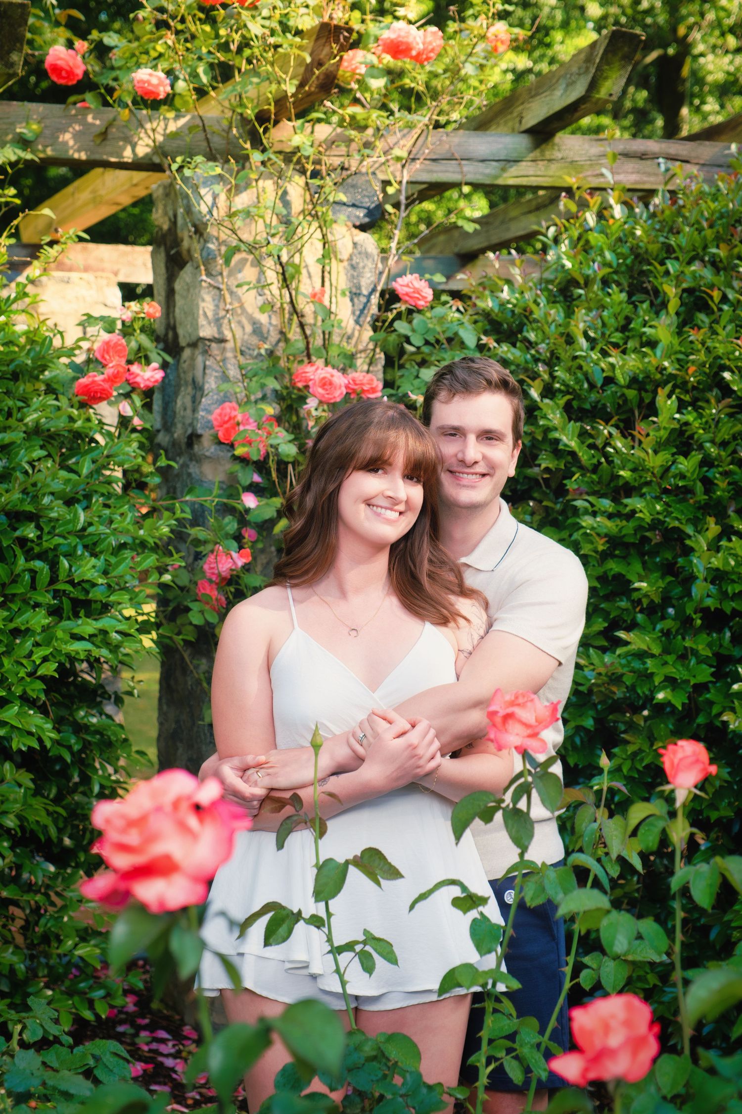 A couple shares a romantic moment together in a lush garden setting surrounded by blooming pink roses and greenery.