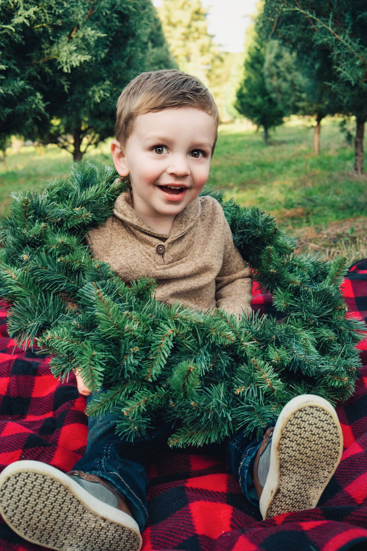 Smiling child sits on plaid blanket with Christmas wreath at tree farm.