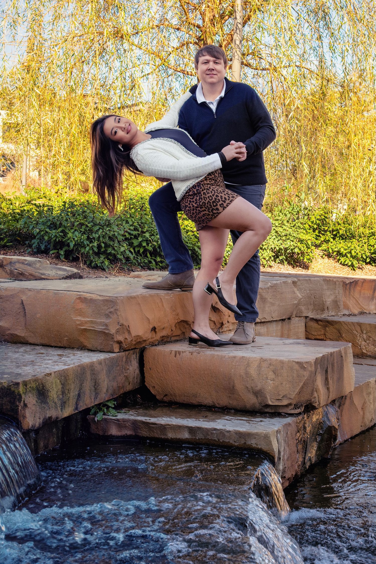 A playful couple poses on stone steps near flowing water with fall foliage in the background.