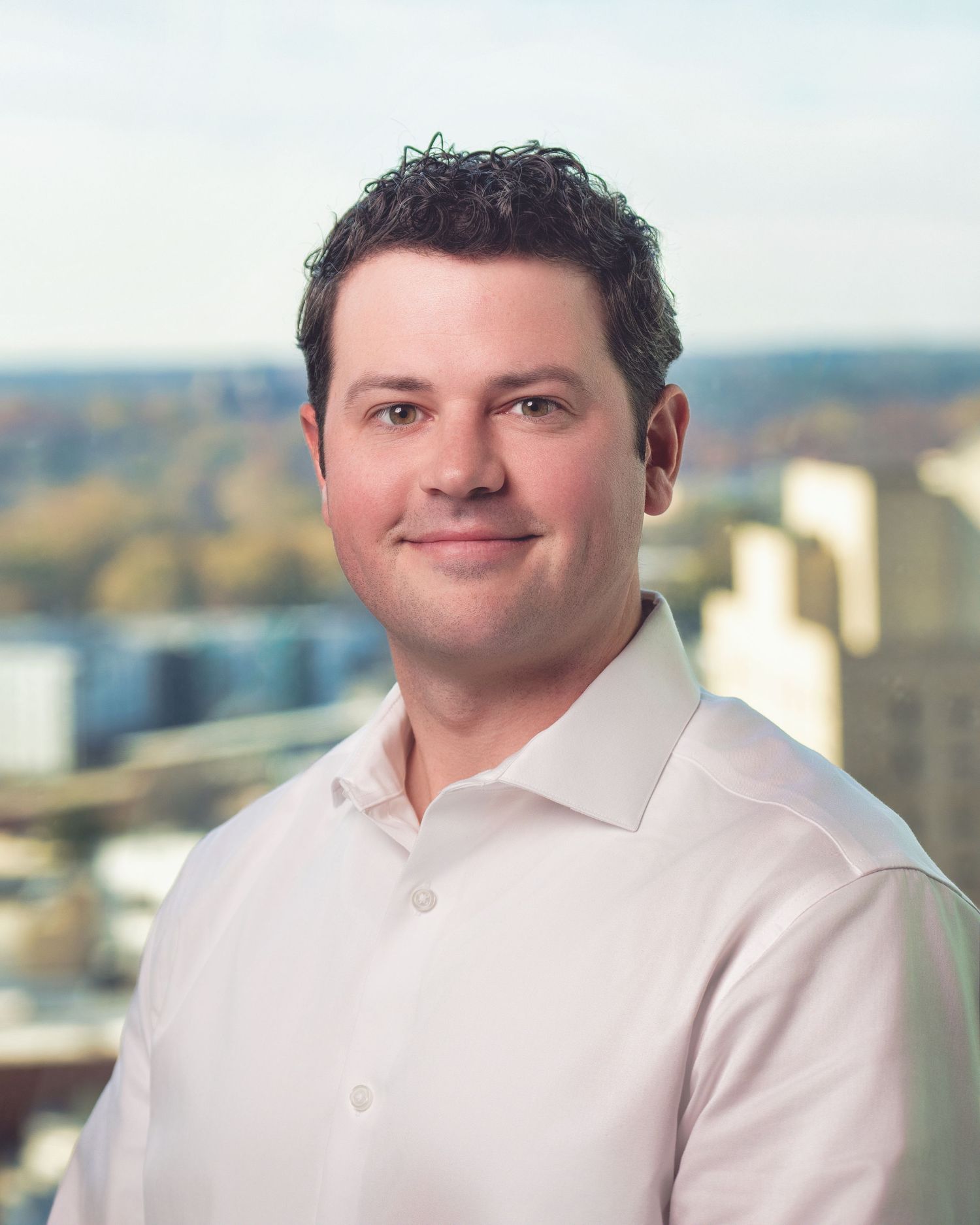 Professional business portrait of person in light pink dress shirt against city skyline background.