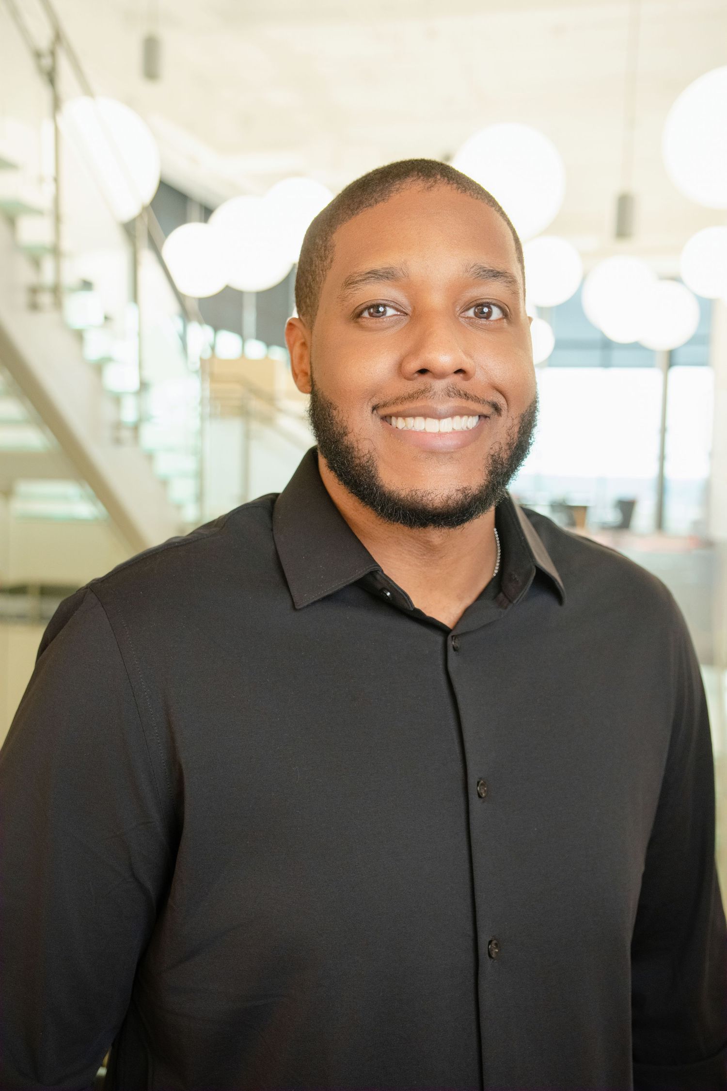 Professional headshot of person in black button-up shirt smiling warmly in bright modern office setting.