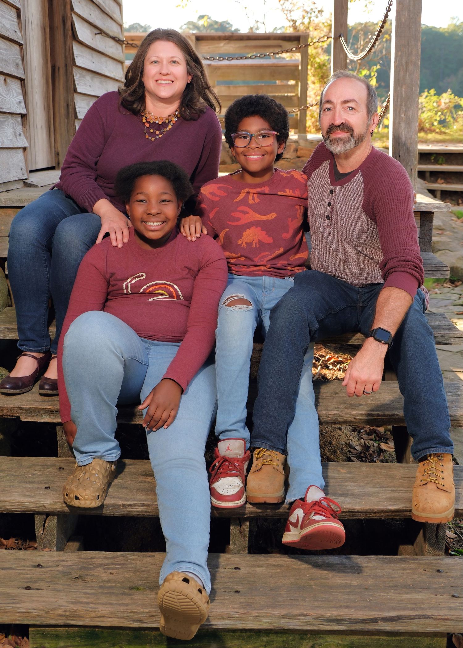 Family of four sitting together on wooden porch steps wearing coordinating burgundy outfits.