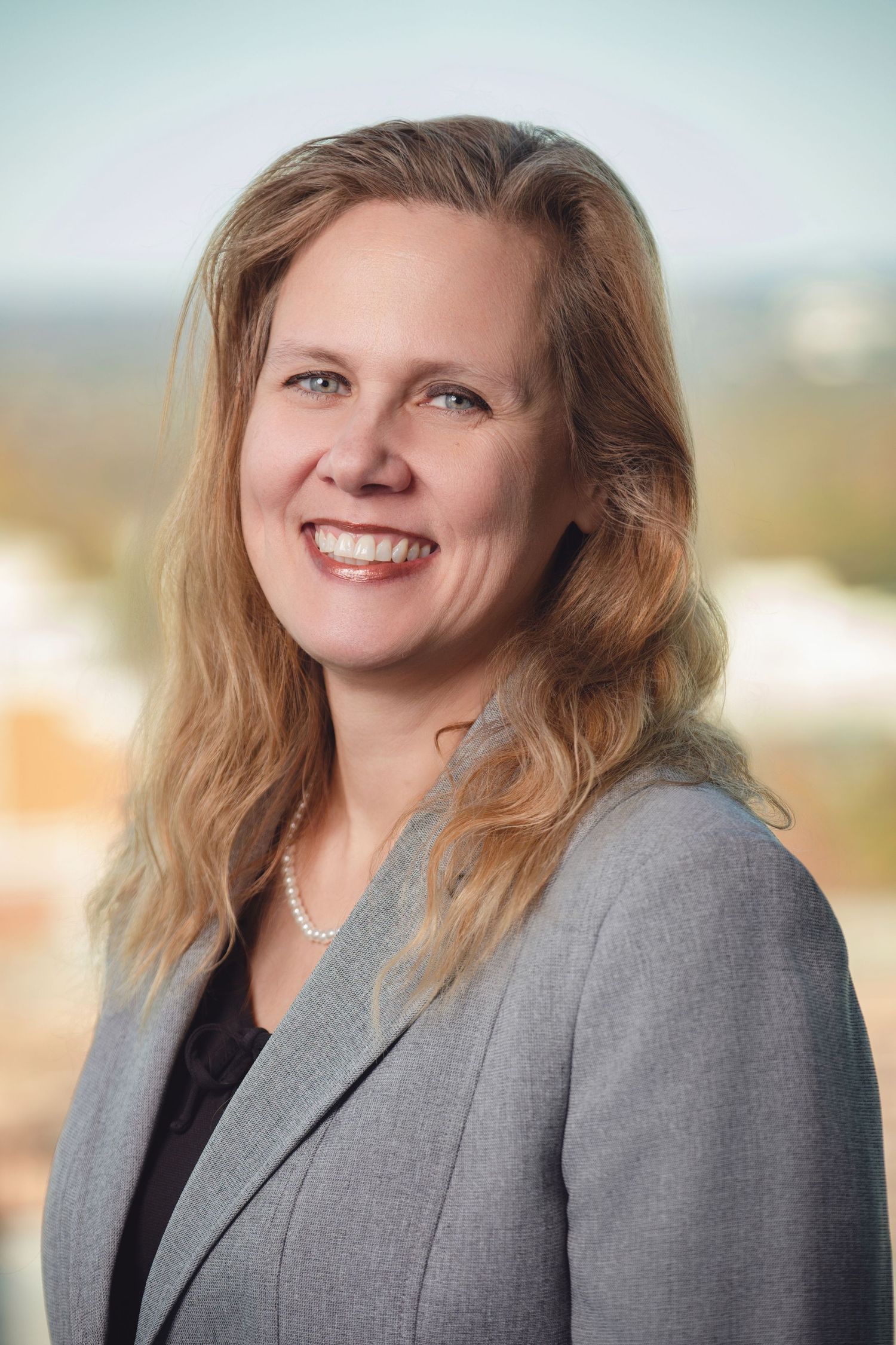 Professional headshot of person in grey suit jacket with wavy hair against soft background.
