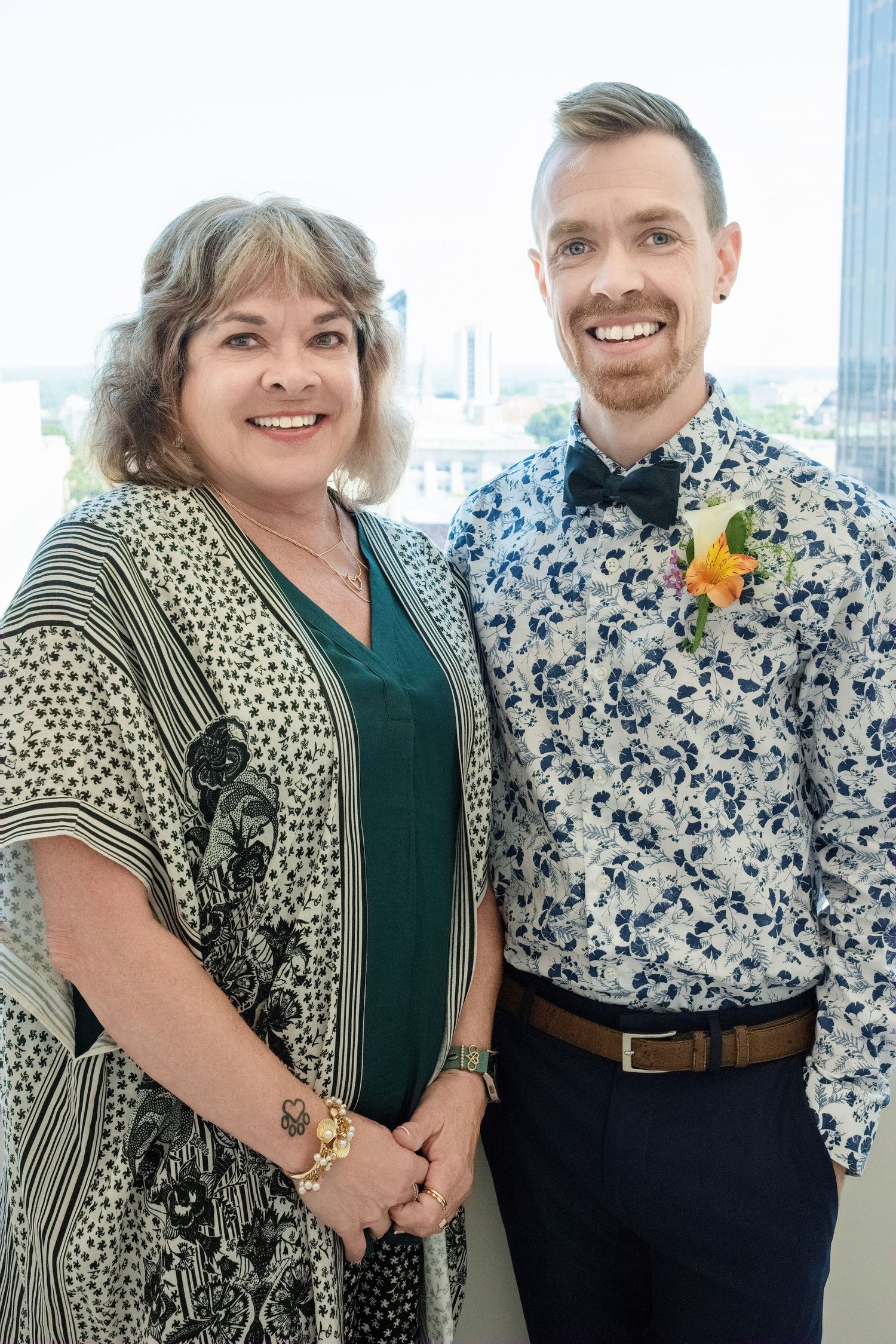 Two people stand close together smiling for a photo, wearing a patterned dress and floral print shirt with a yellow boutonniere.