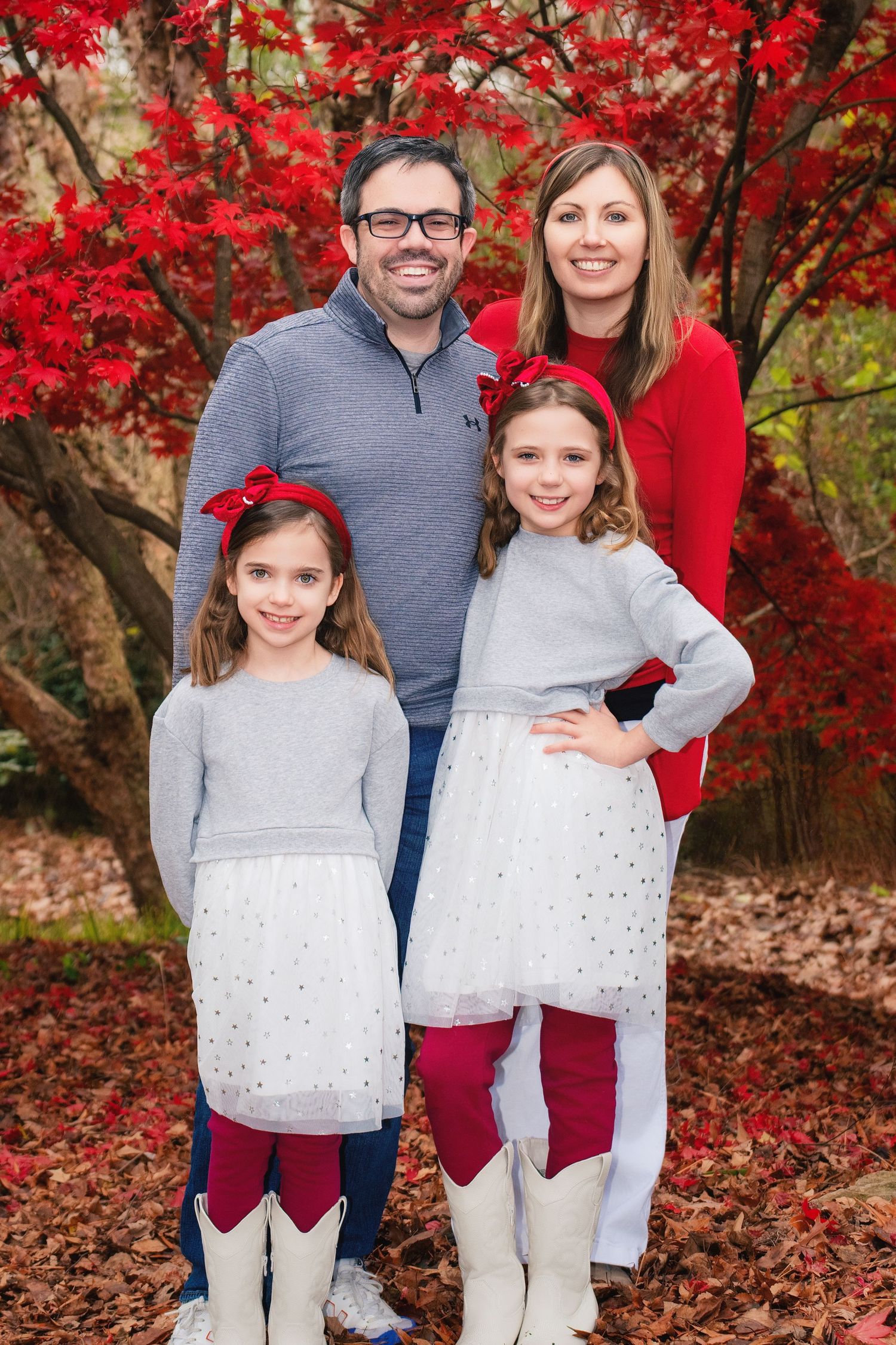 A family poses together outdoors in coordinated outfits against vibrant red autumn foliage and fallen leaves.