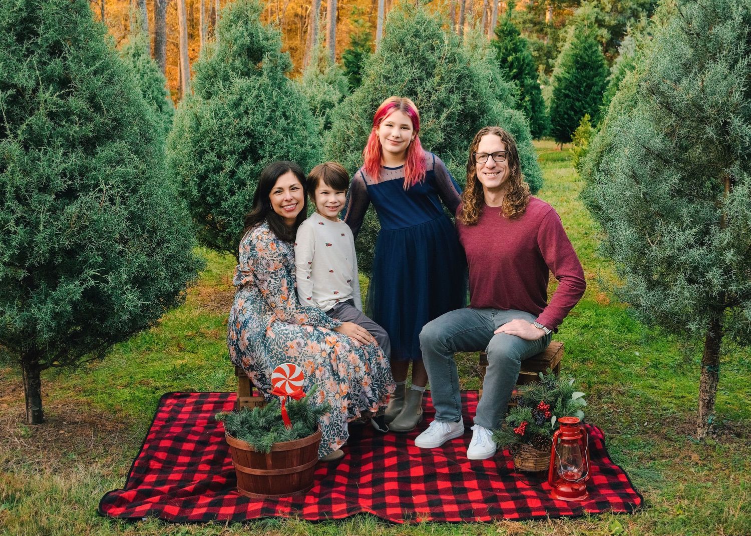 A group poses on a red plaid blanket among Christmas trees at sunset.