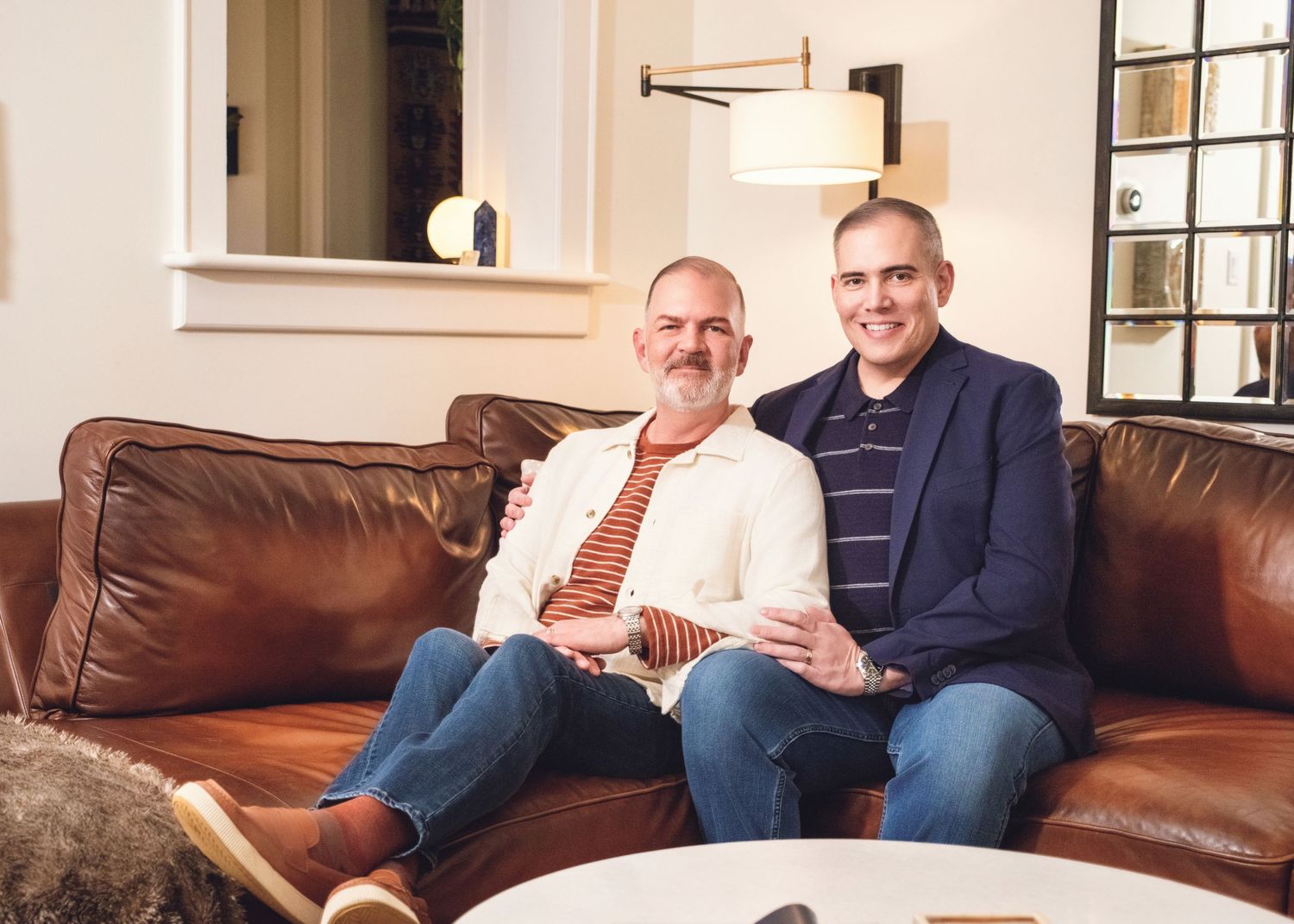 Two men sit close together on a brown leather couch in a bright living room interior.