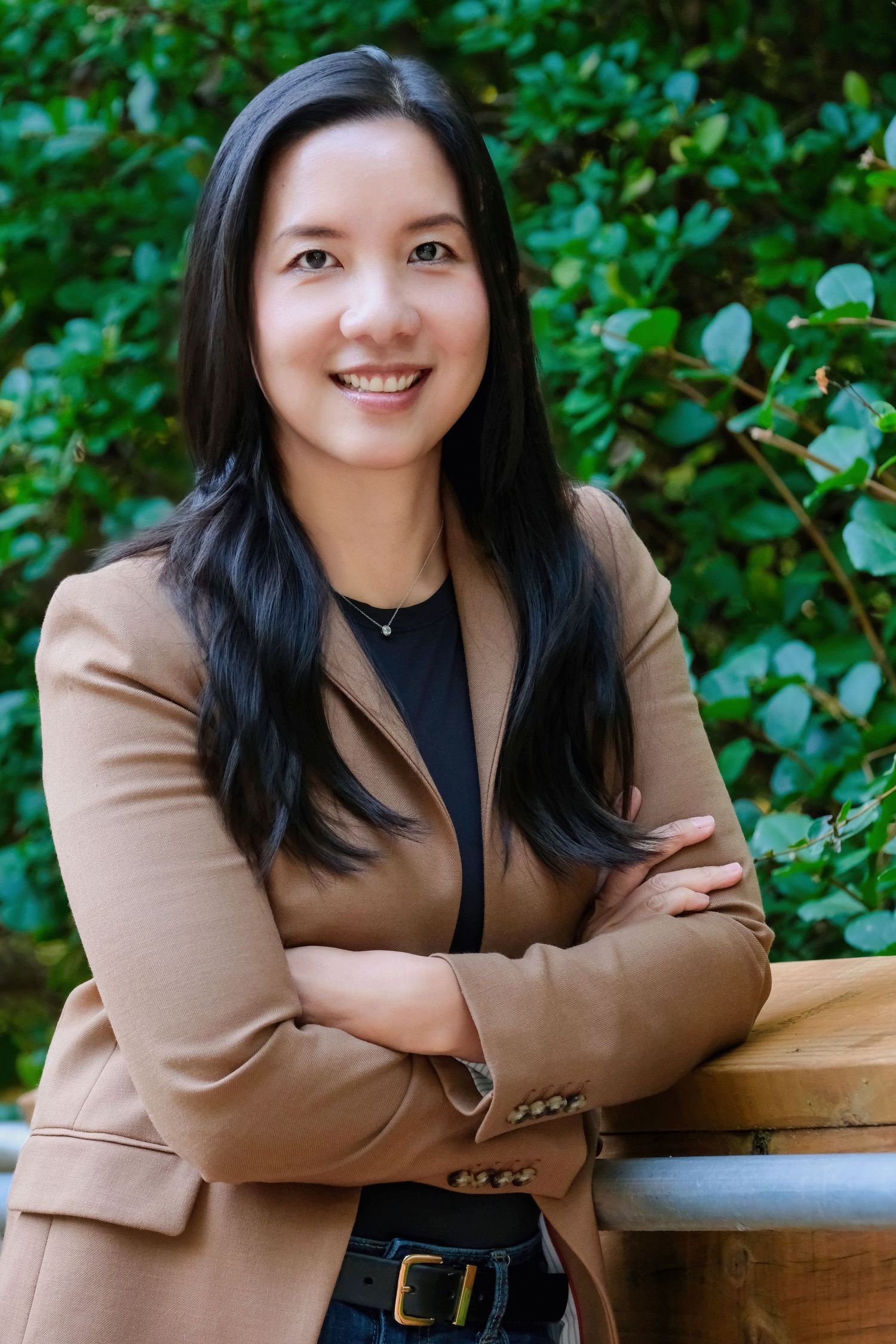 Professional portrait of someone in a tan blazer posing against green foliage background.