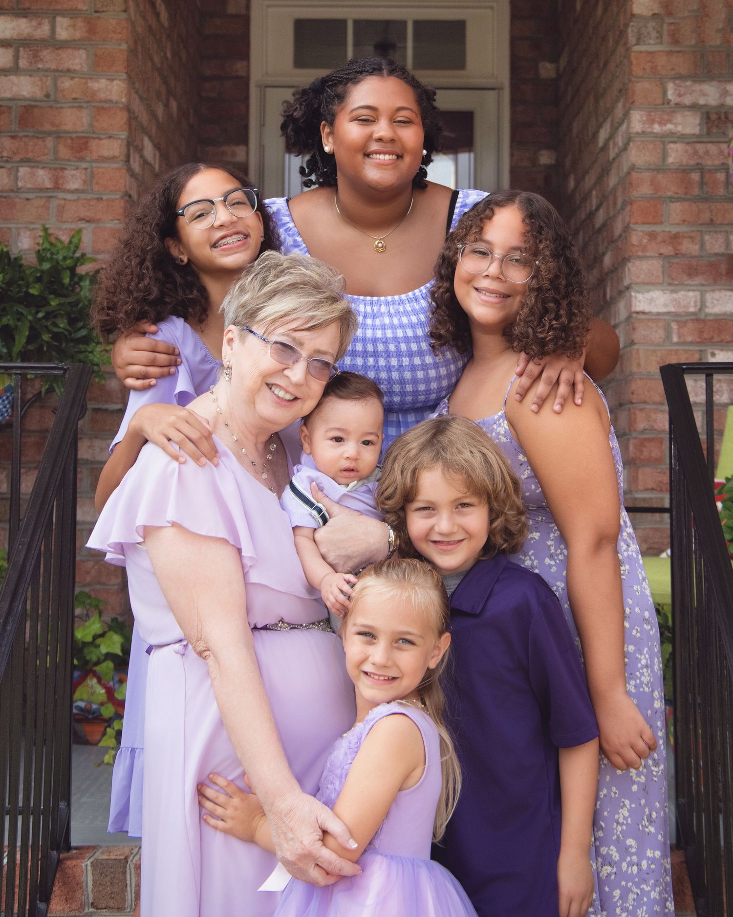 A multi-generational family wearing coordinated purple and pink outfits poses together on a brick porch.