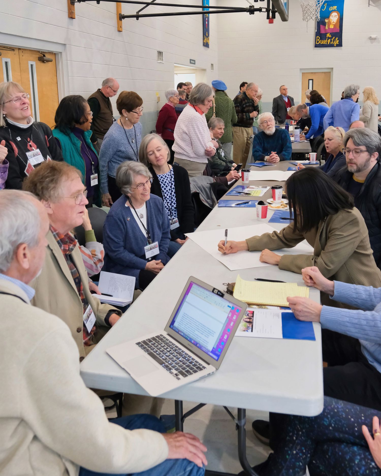 People gathered at tables in a community meeting or registration event with laptops and paperwork in a gymnasium space.