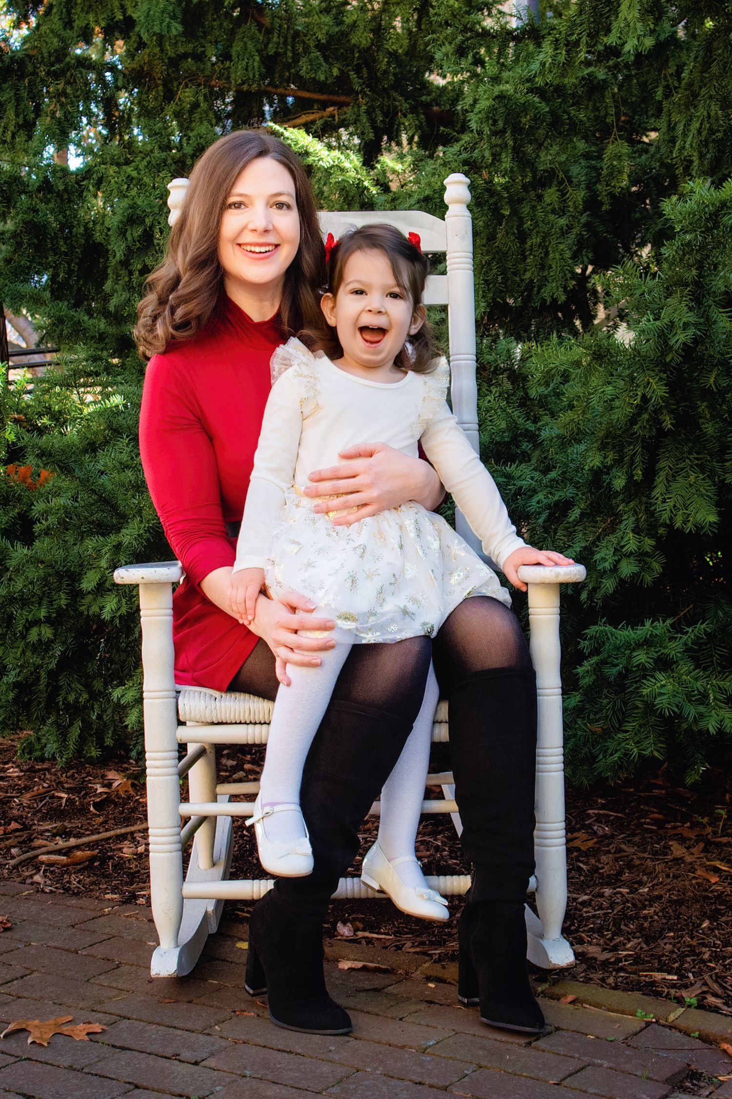 A child and adult sit together in white rocking chairs on a wooden deck surrounded by lush greenery.