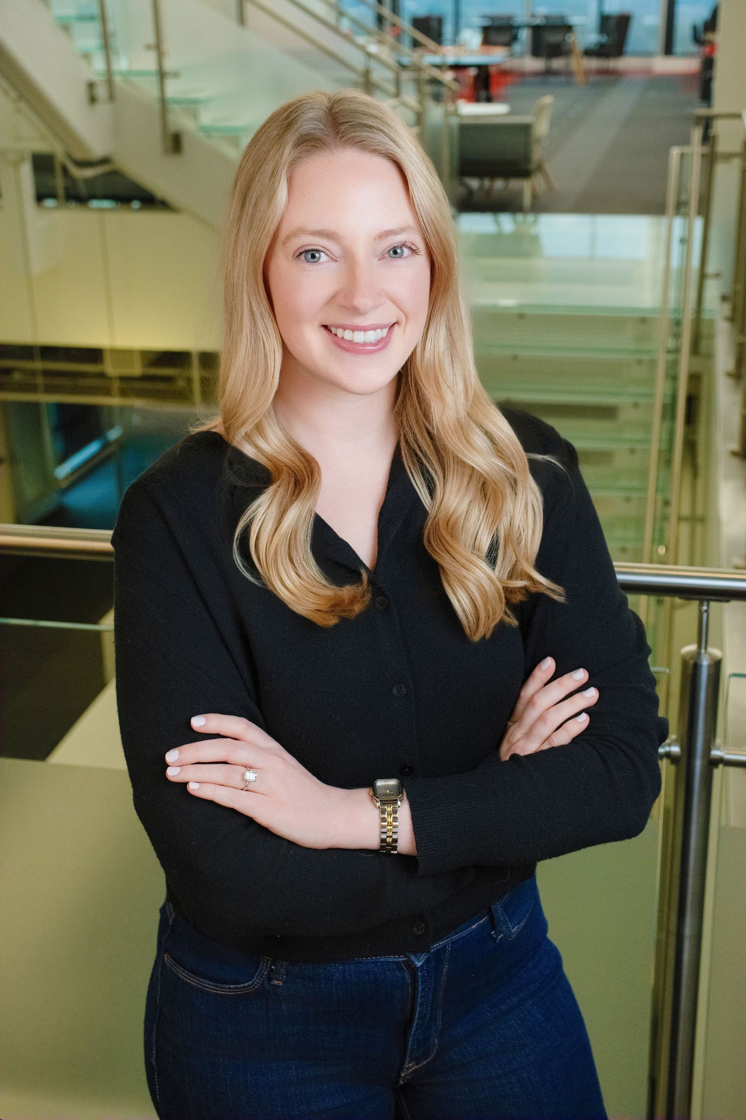 Professional headshot of person in black sweater and jeans standing with arms crossed in modern office setting.