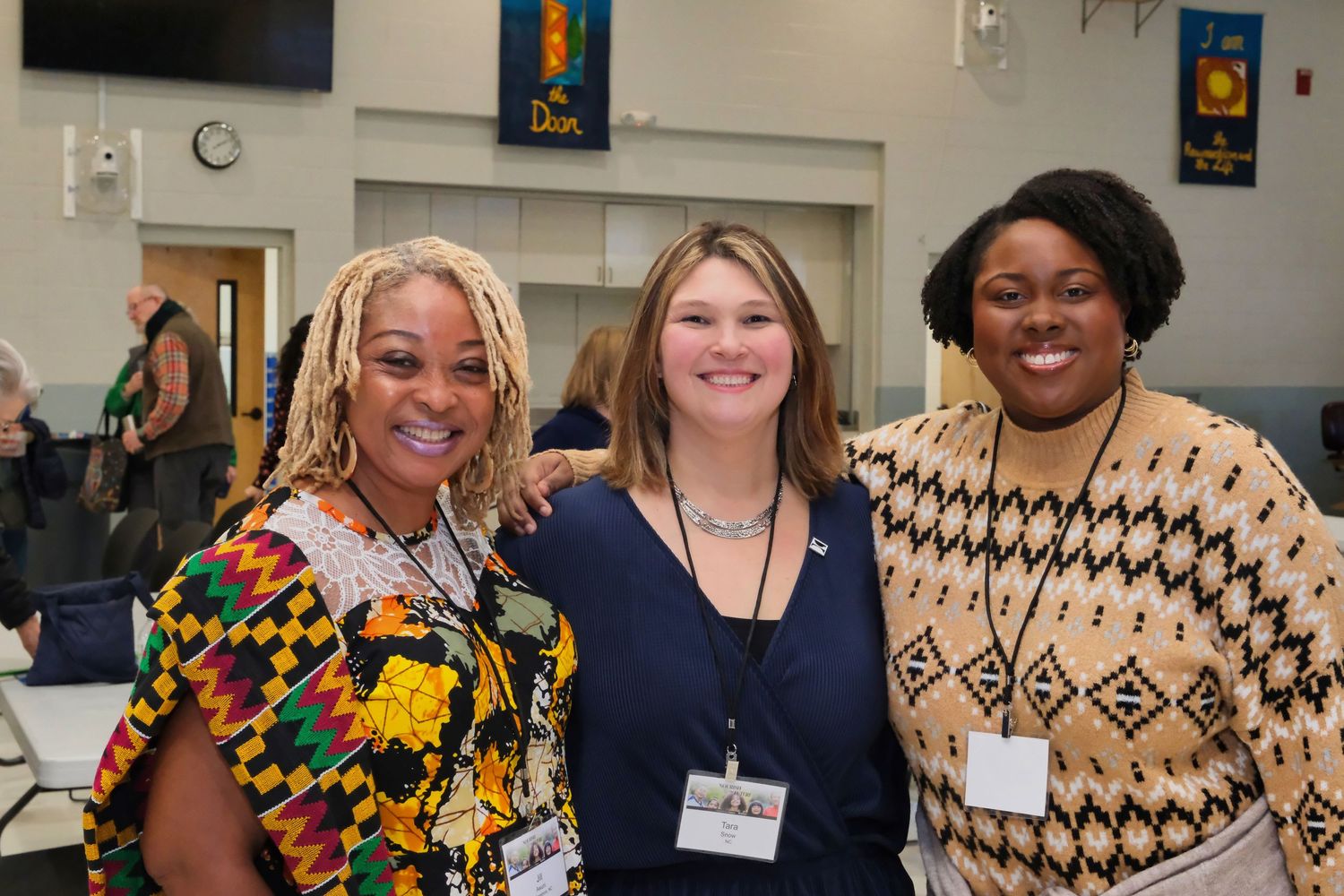 Three people smiling together at a community event, wearing colorful outfits and name badges.