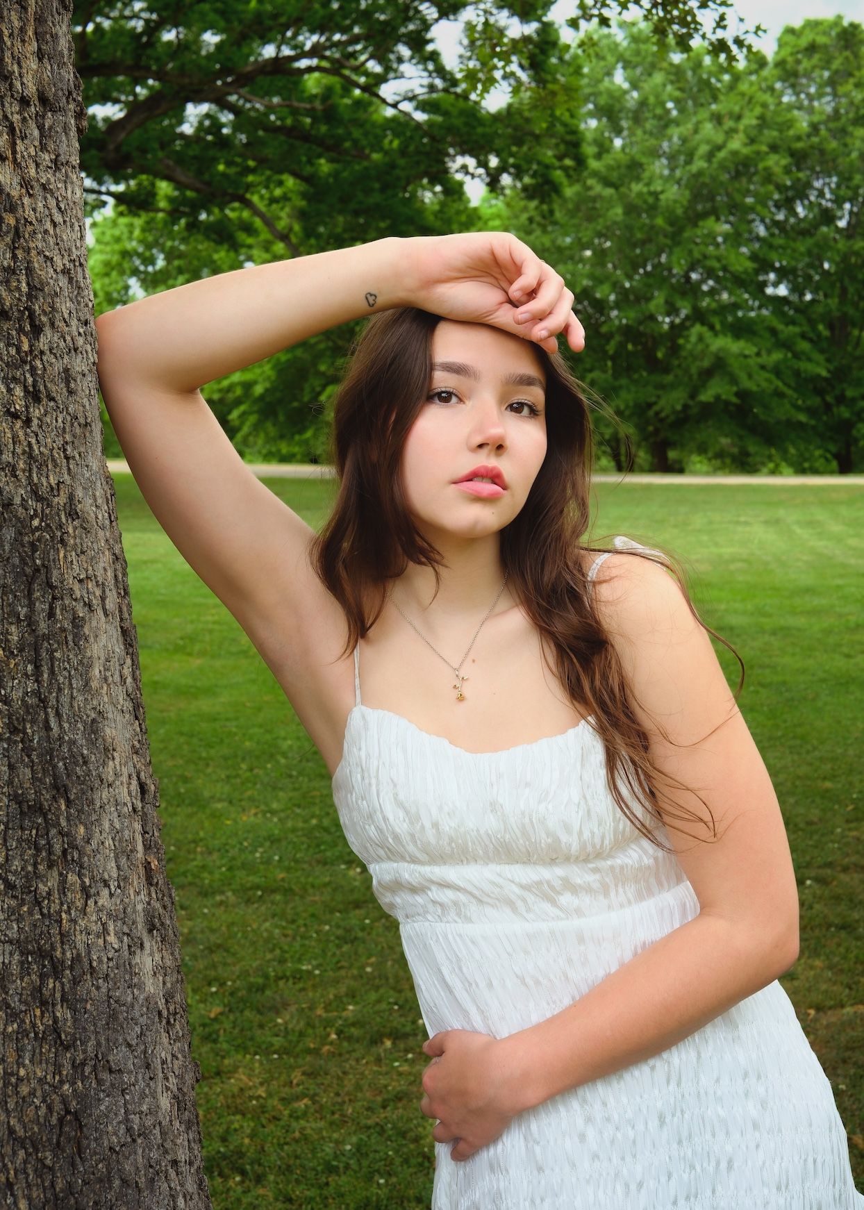Person in white dress poses elegantly near tree in green park.