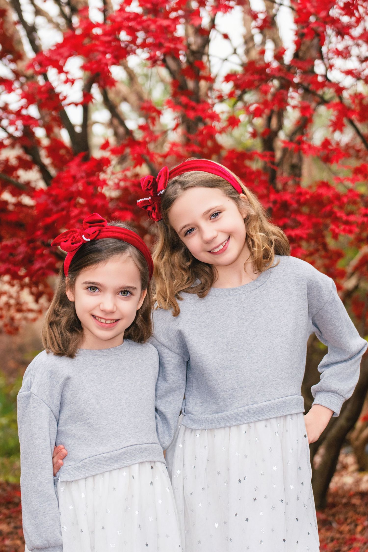 Two figures in matching gray sweaters pose together against a backdrop of vibrant red autumn foliage.